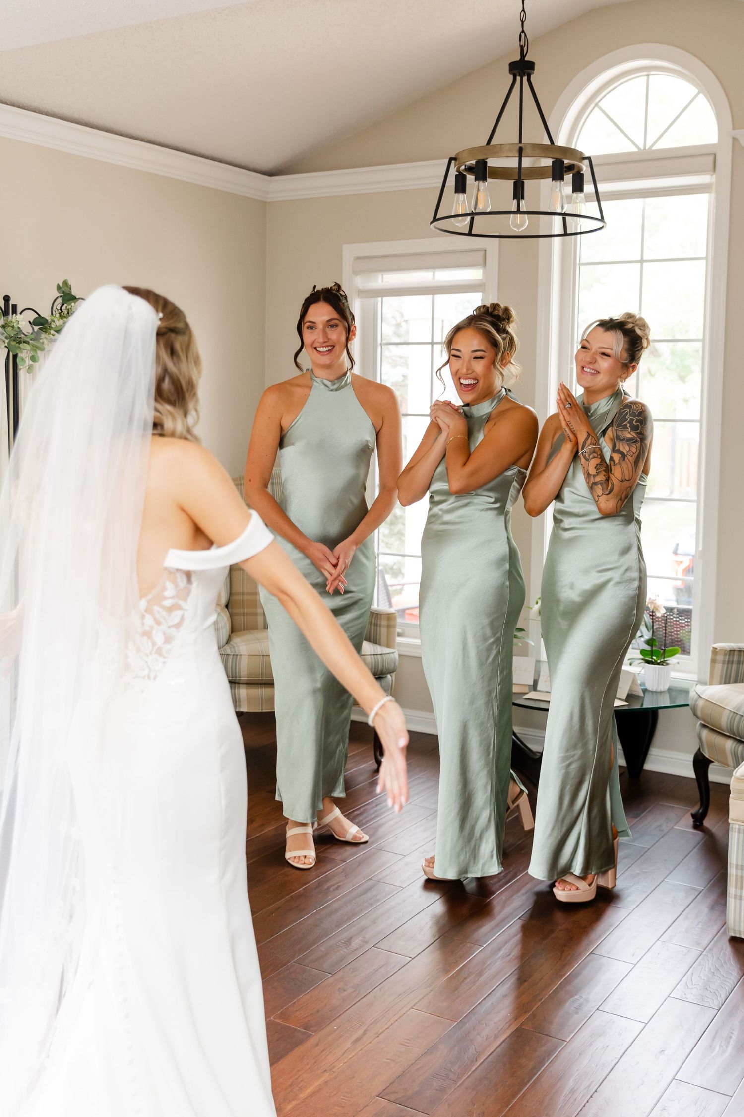 Bridesmaids in sage green dresses share excited reactions during a wedding day reveal.