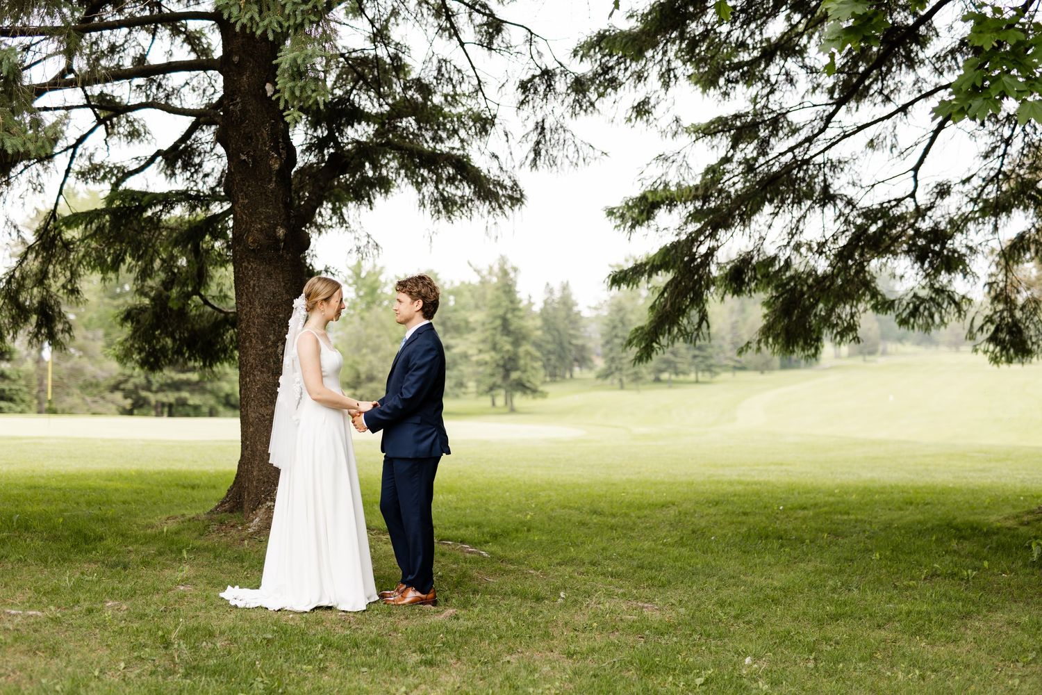 A wedding couple stands together on a serene golf course with evergreen trees in the background.