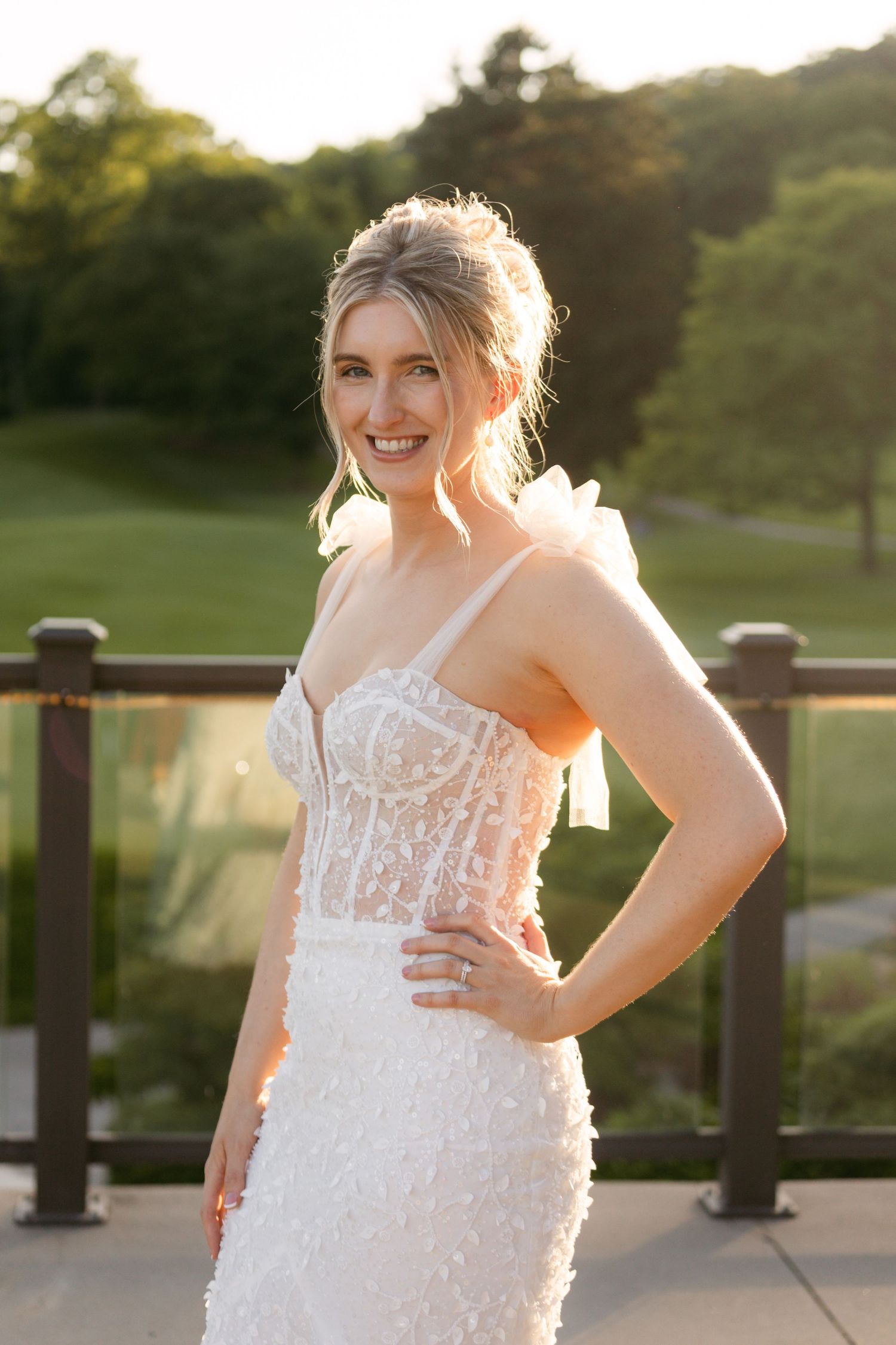 Bride in a white lace gown poses joyfully on an outdoor terrace during sunset.