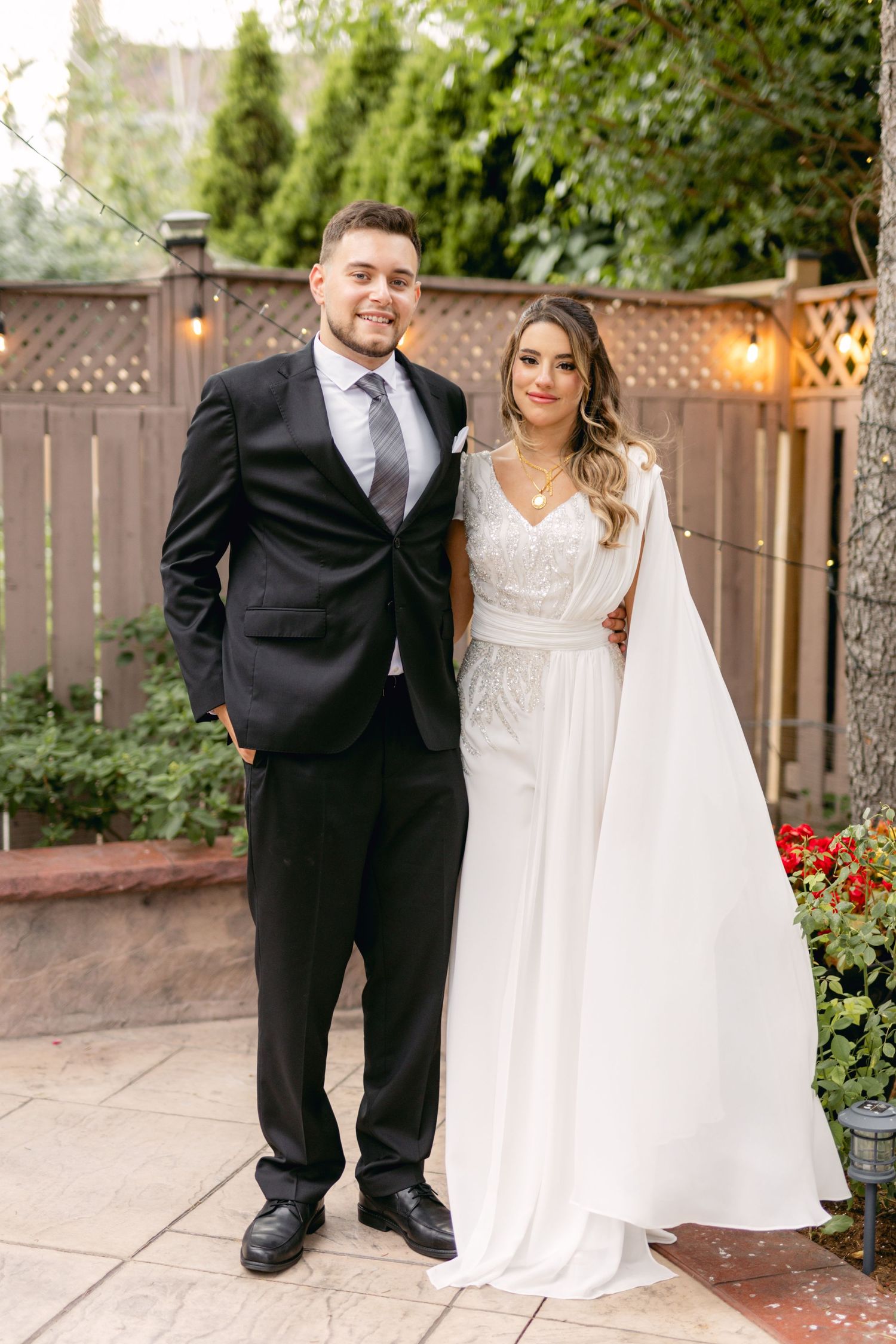 Wedding couple poses in garden setting with wooden fence and string lights.