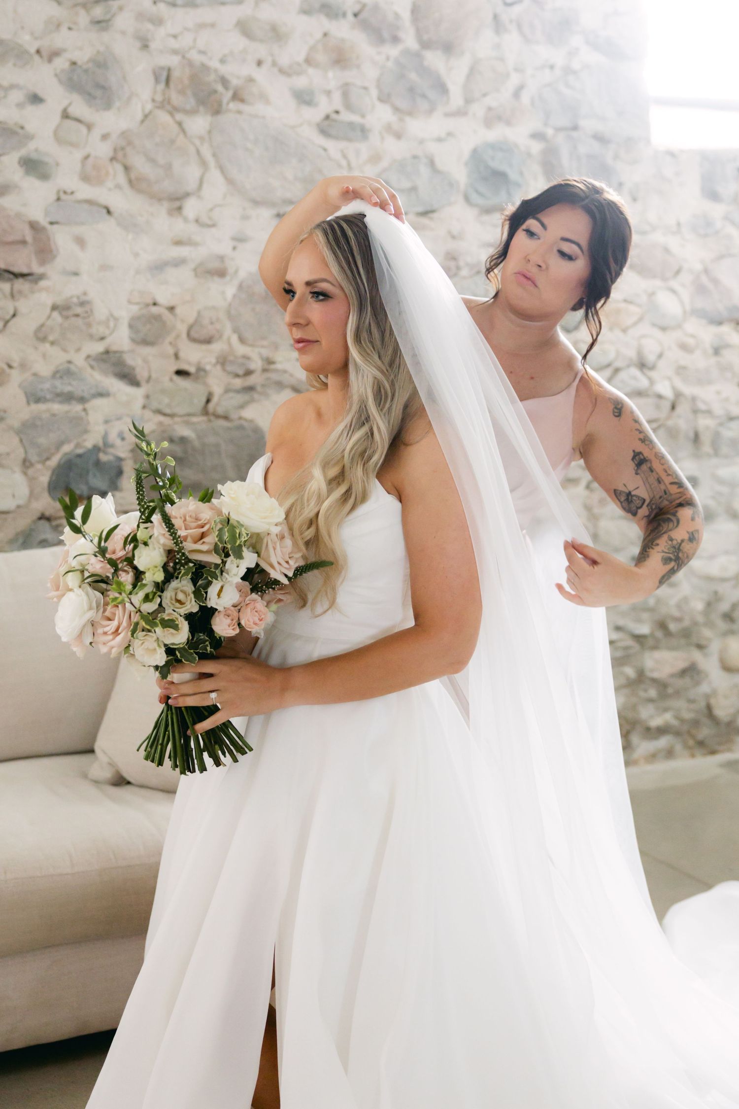 Wedding preparation moment with bride holding bouquet of pink and white flowers against stone wall.