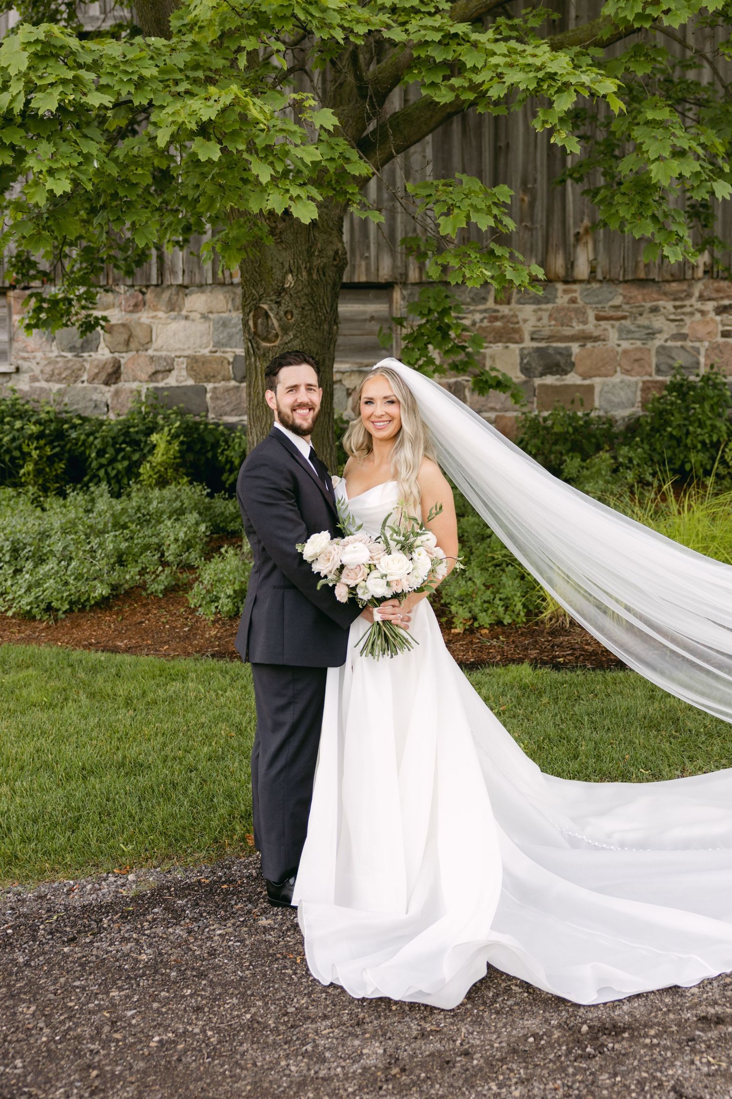 Bride and groom pose with dramatic flowing veil against stone wall and greenery.