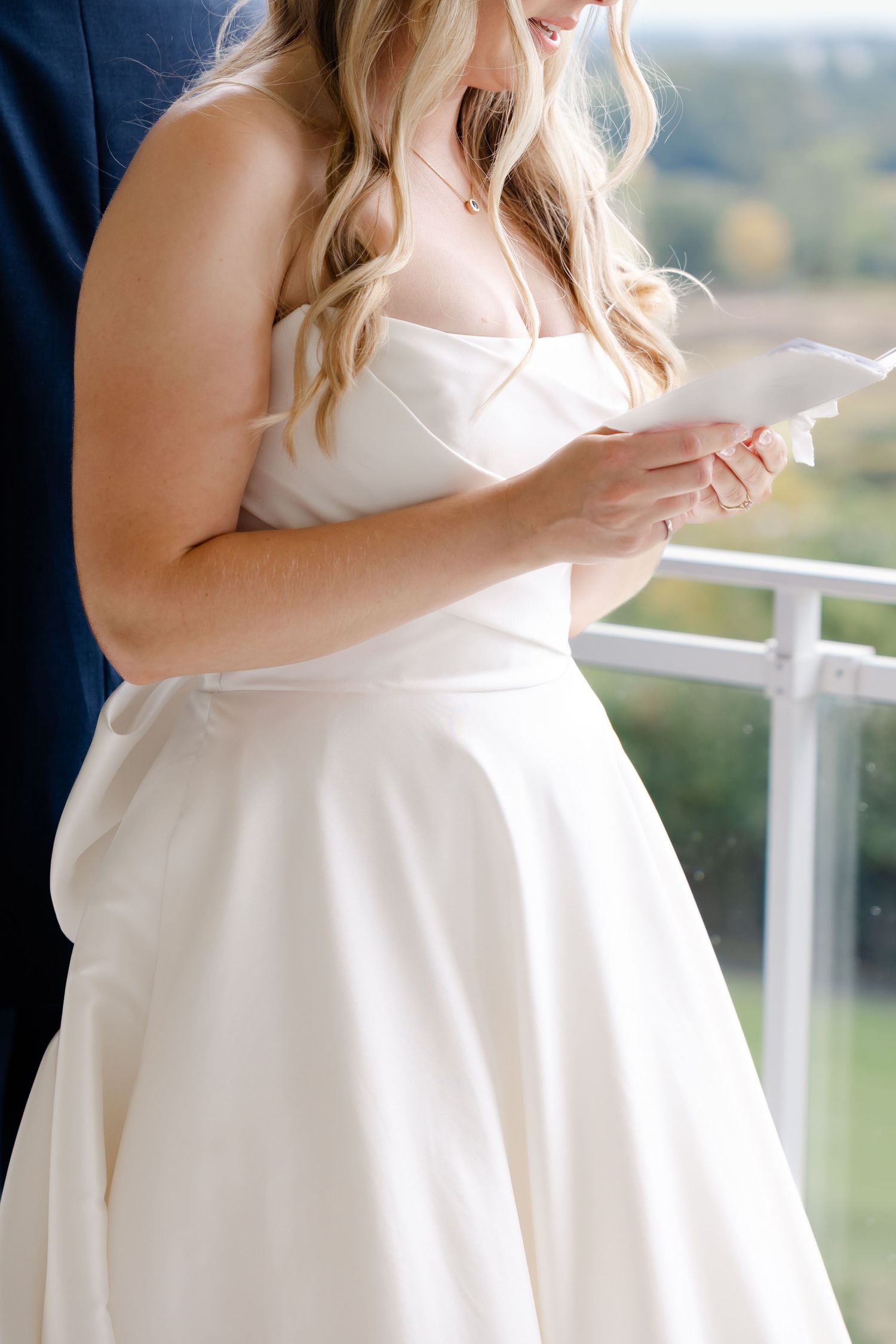 Close-up of a bride in a white wedding dress reading a note on a balcony.
