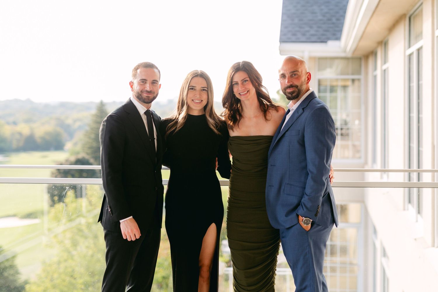 Two couples dressed formally pose together on an outdoor balcony with scenic views.