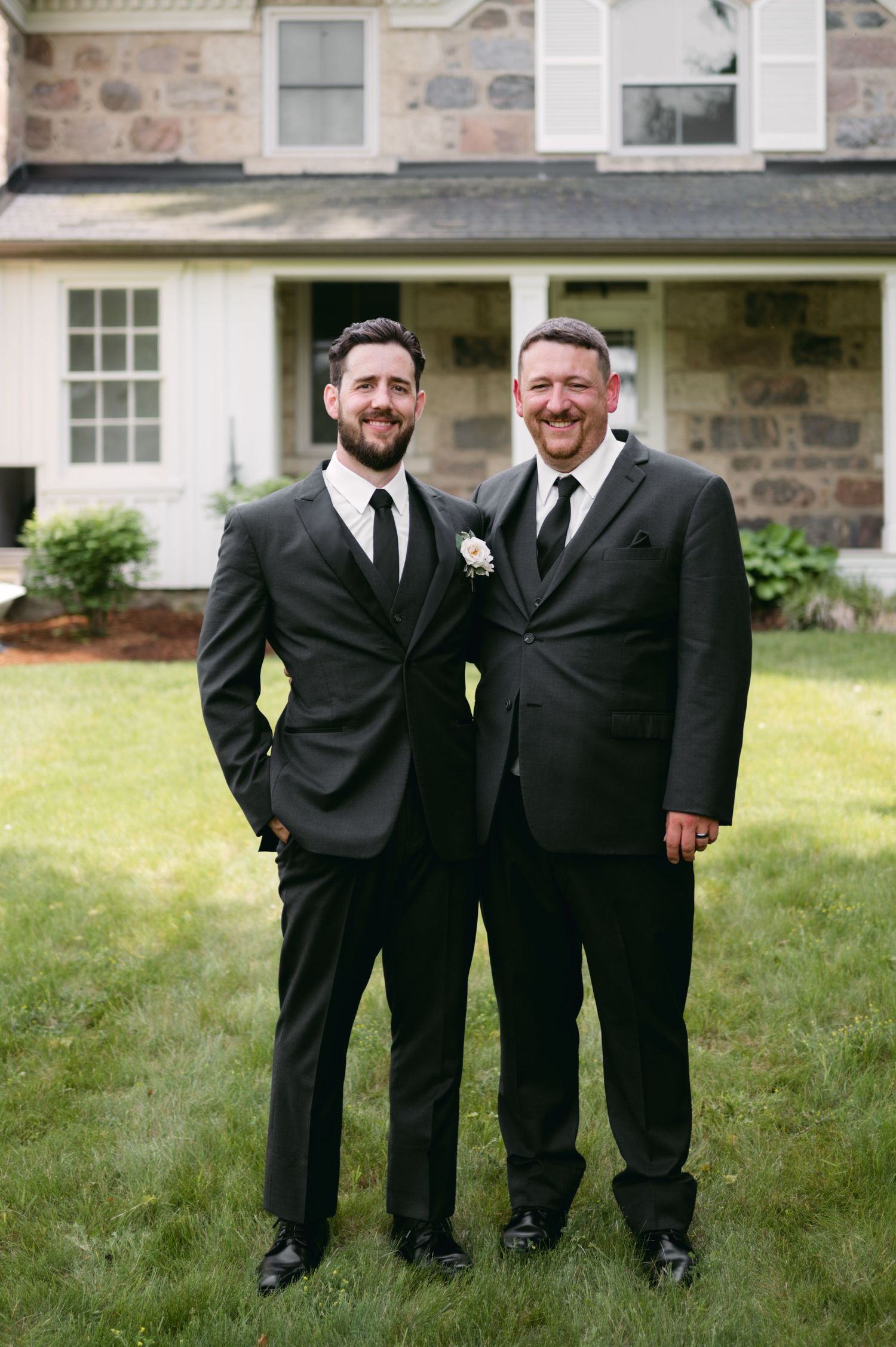 Groomsmen in classic black suits pose casually on lawn near stone building.