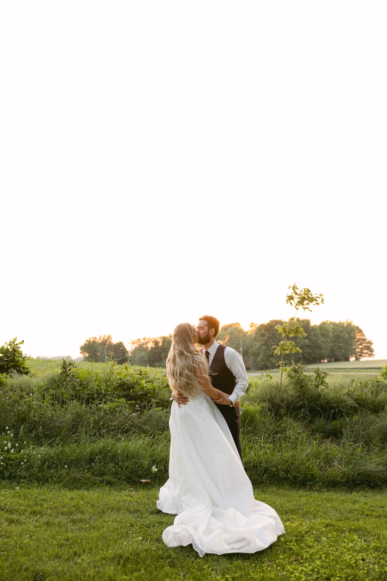 Bride and groom dance romantically in a lush green field during golden hour.