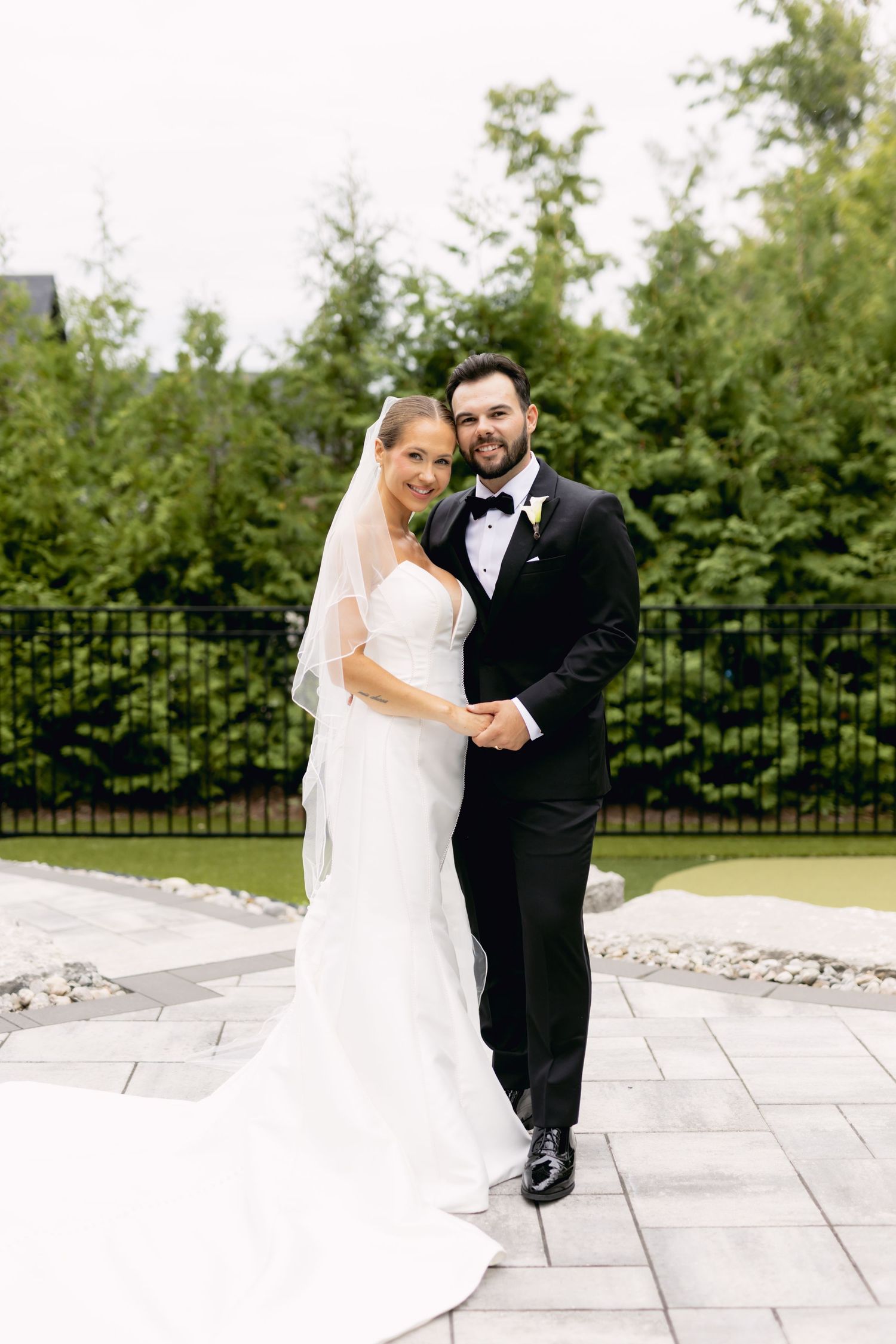 Smiling bride and groom pose together on stone patio, bride in white mermaid gown with veil, groom in black tux.