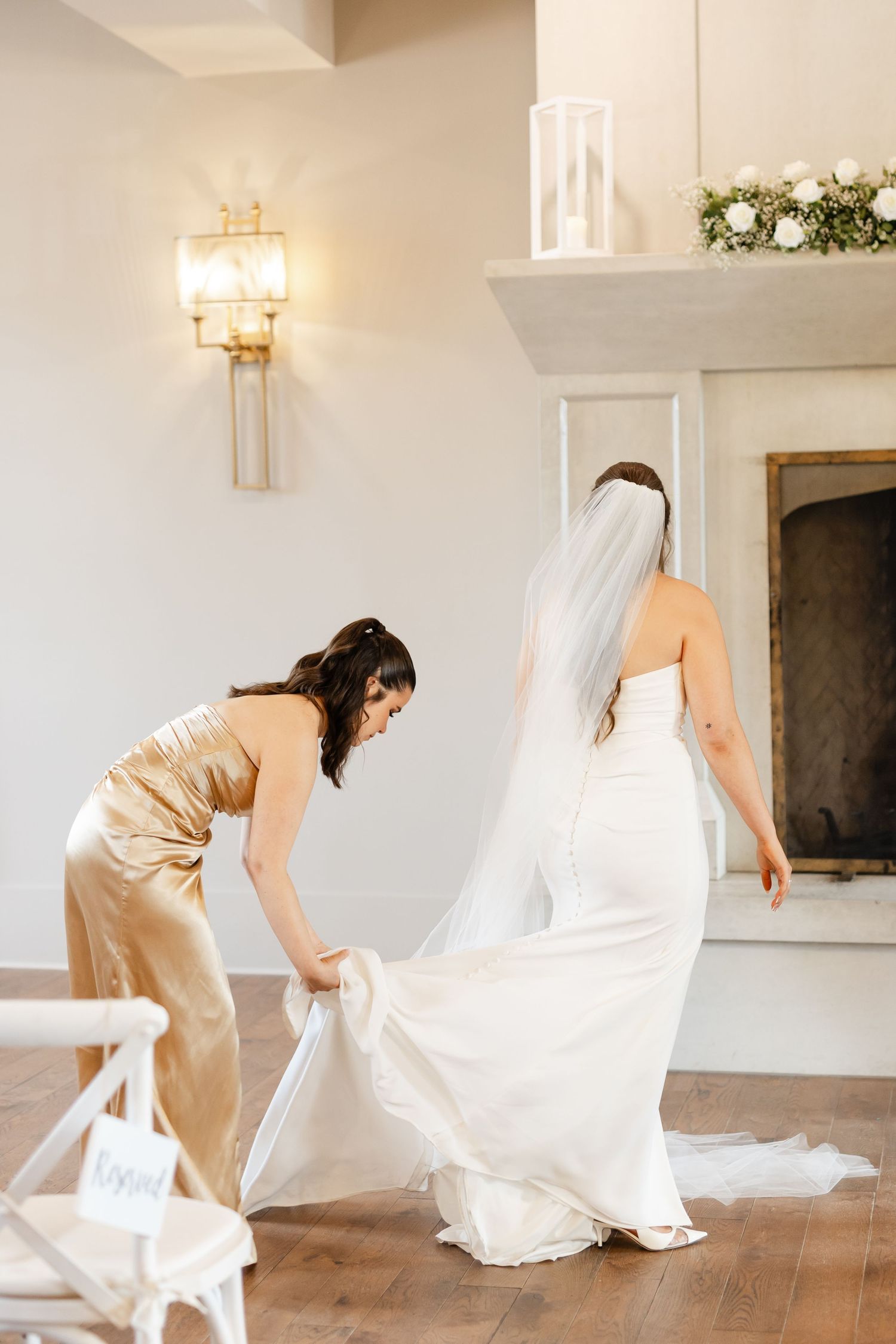 Bridesmaid in gold dress adjusts bride's cathedral veil near fireplace before wedding ceremony.