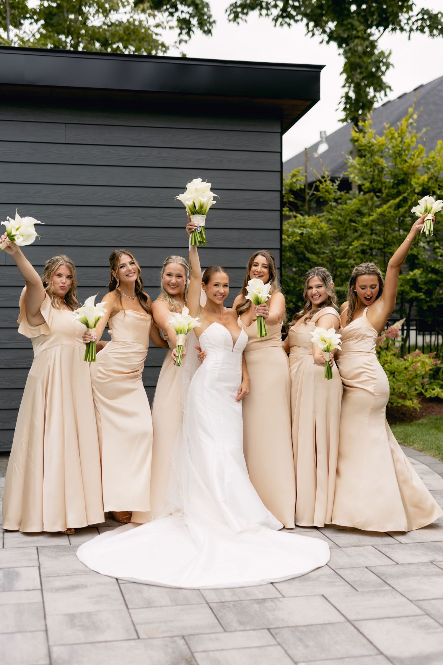 Bride in white gown poses with six bridesmaids in champagne dresses holding white bouquets outdoors.