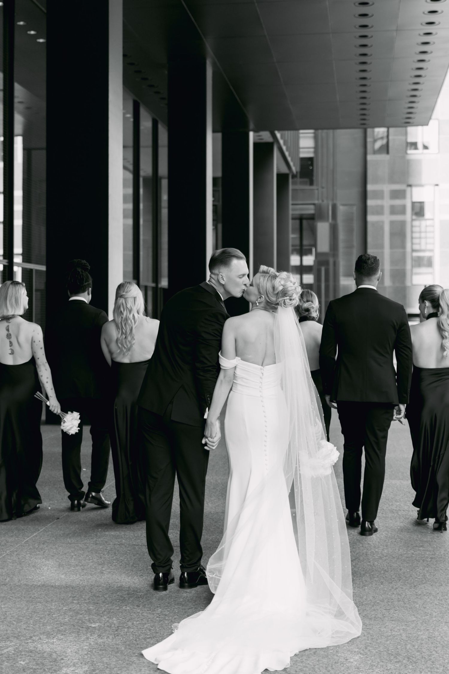 Black and white photo of bride and groom holding hands with wedding party walking ahead in city.
