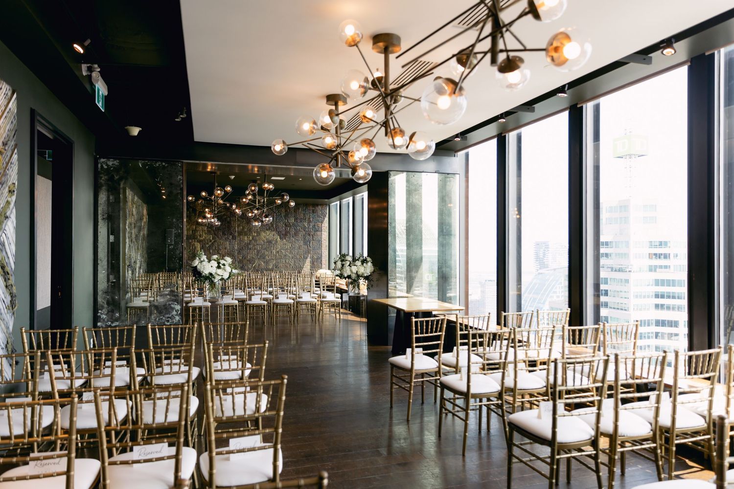 Modern wedding ceremony venue with gold chiavari chairs, chandelier lighting, and floor-to-ceiling city views.