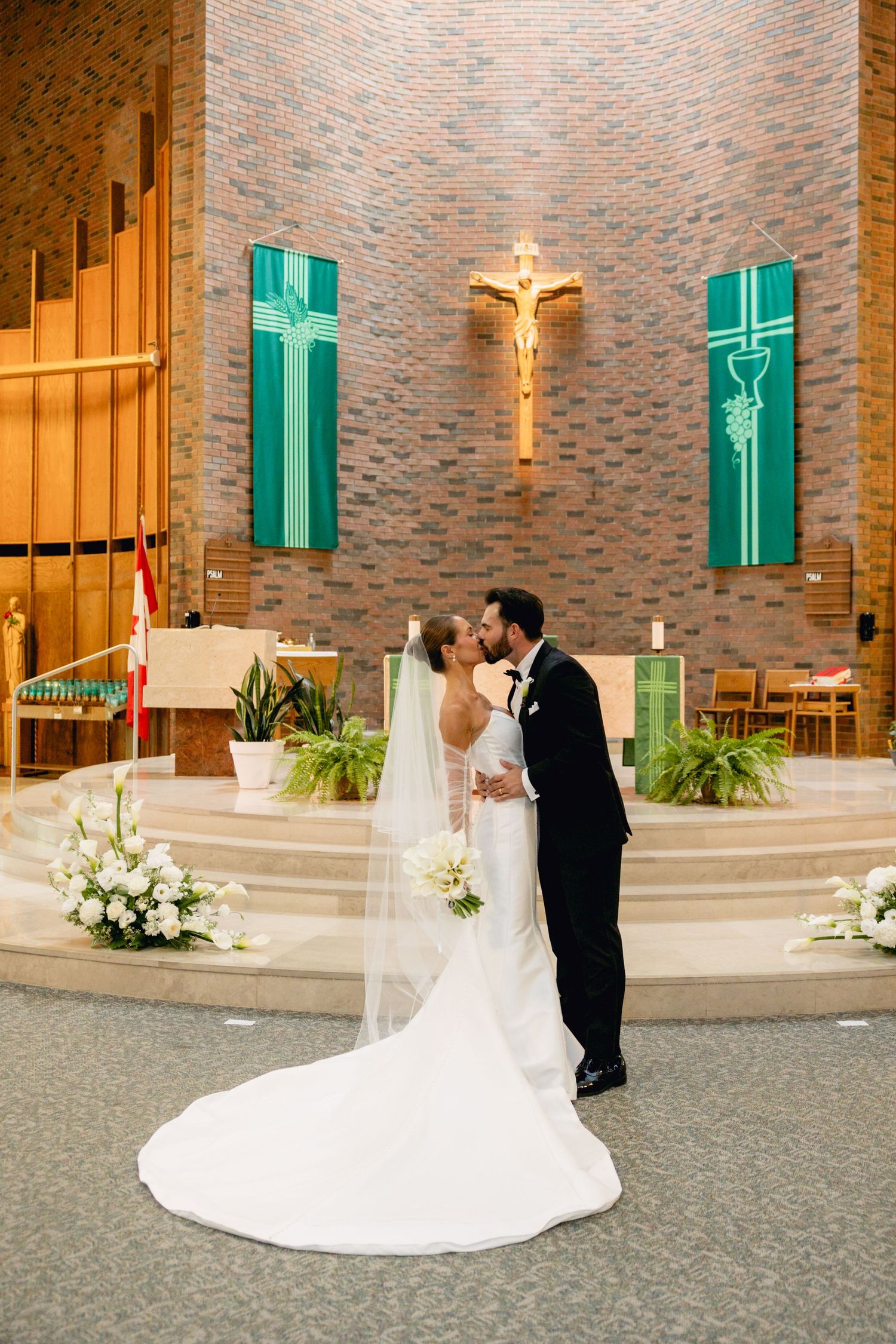Bride and groom sharing first kiss at church altar with crucifix and green banners behind them.