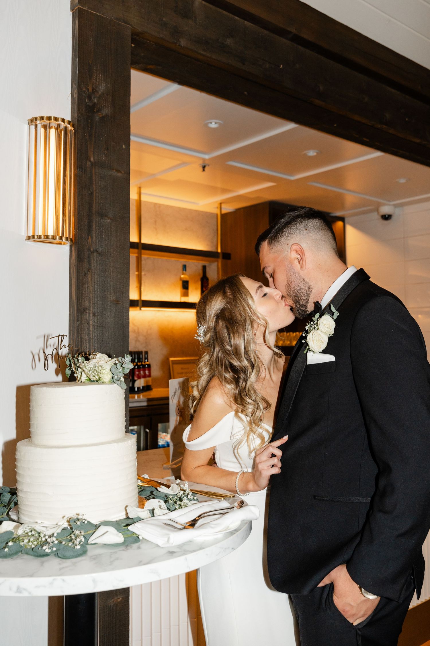Newlyweds share intimate moments while cutting their elegant white wedding cake adorned with greenery.
