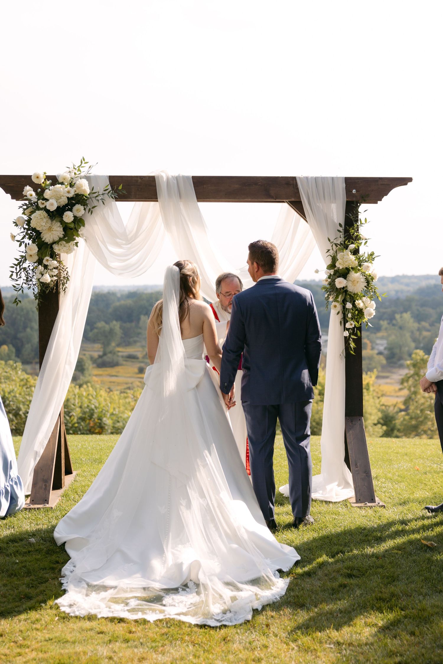 Wedding ceremony under white draped pergola decorated with floral arrangements on a hilltop.