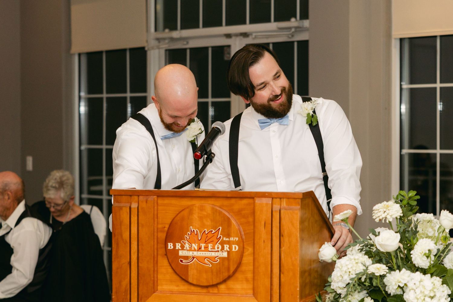 Wedding reception speakers share heartfelt moments at a wooden podium decorated with white floral arrangements.