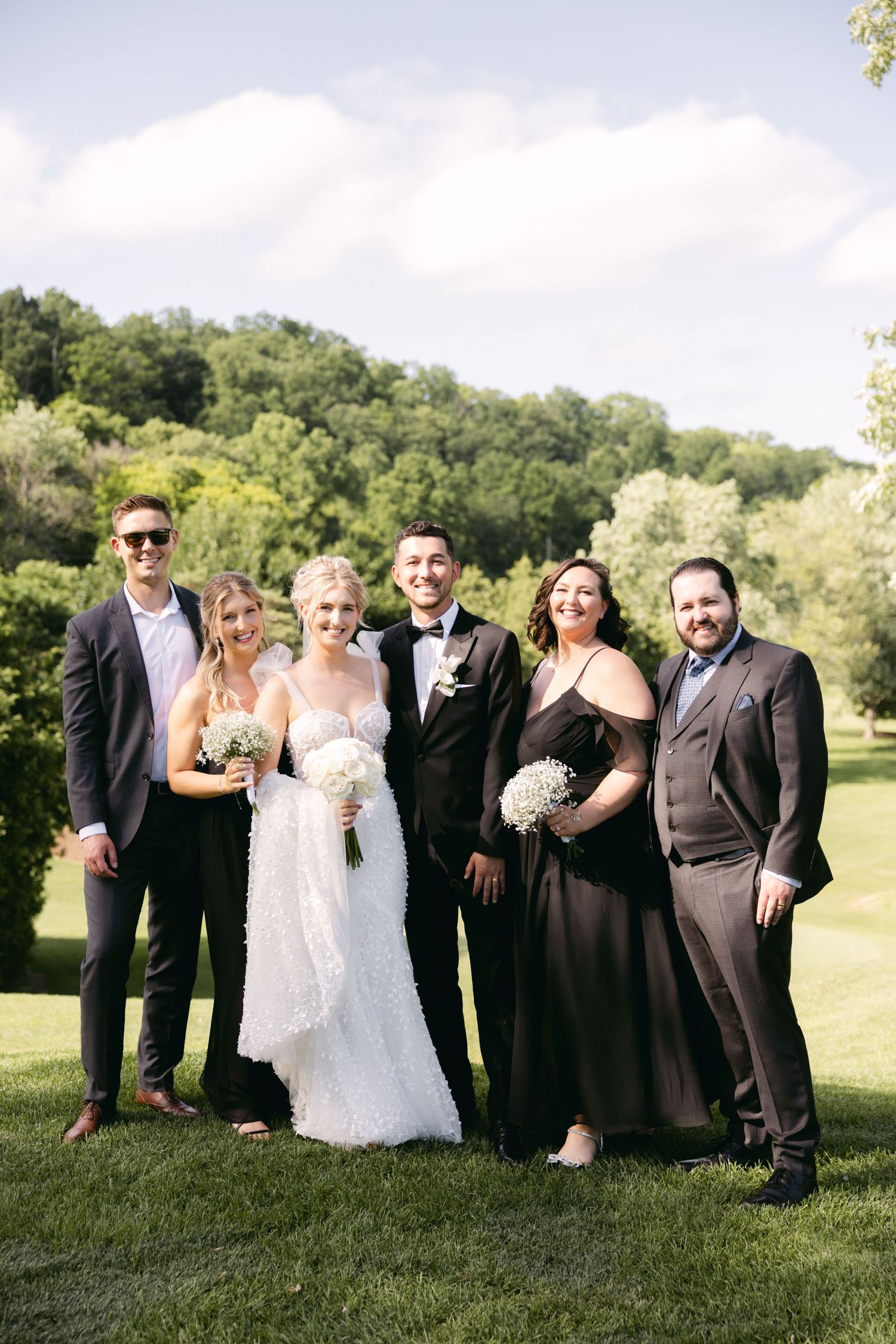 Wedding group gathers for formal portraits on a lush green hillside under summer skies.