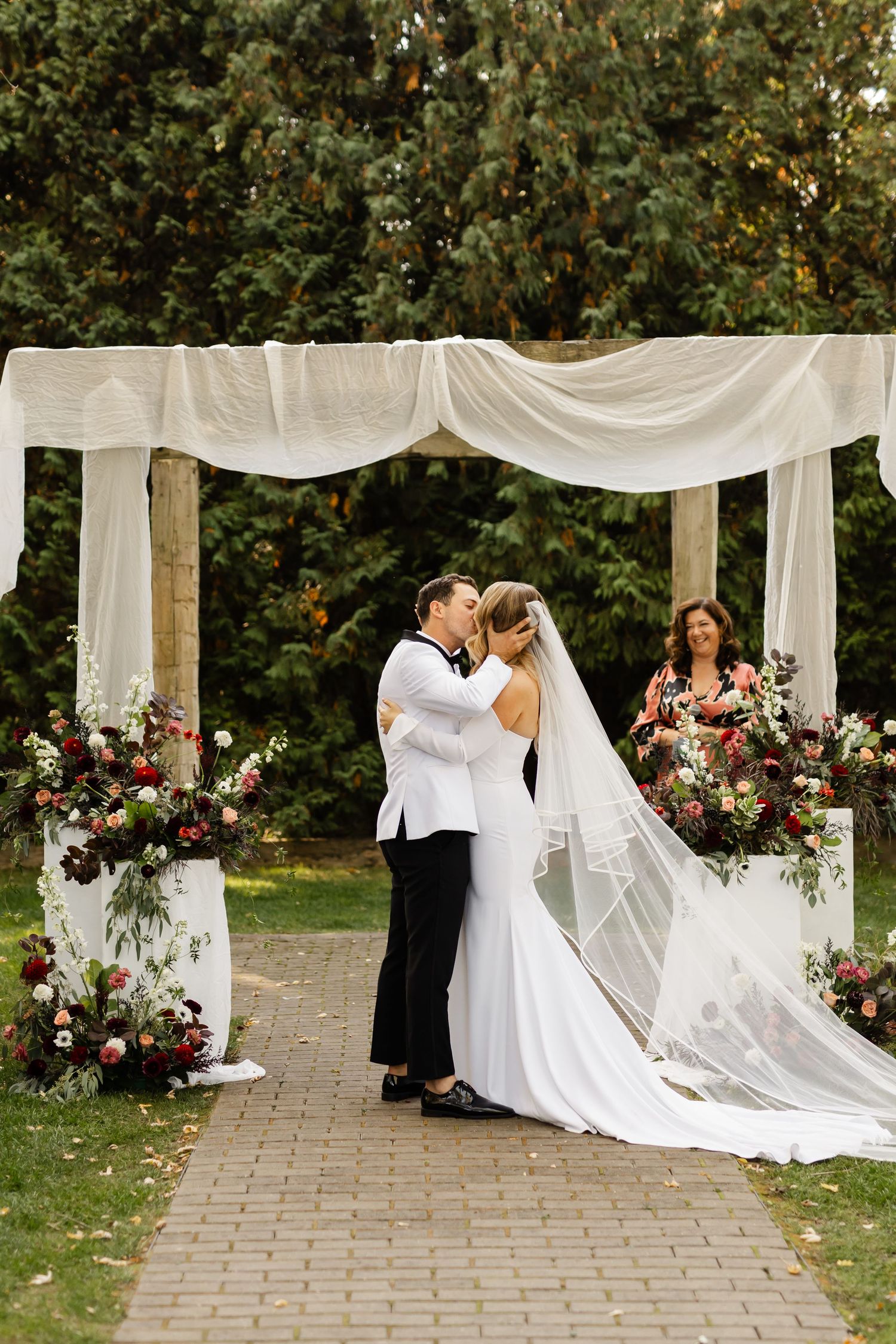 Wedding couple shares intimate moment under white draped arch with bridal party looking on.