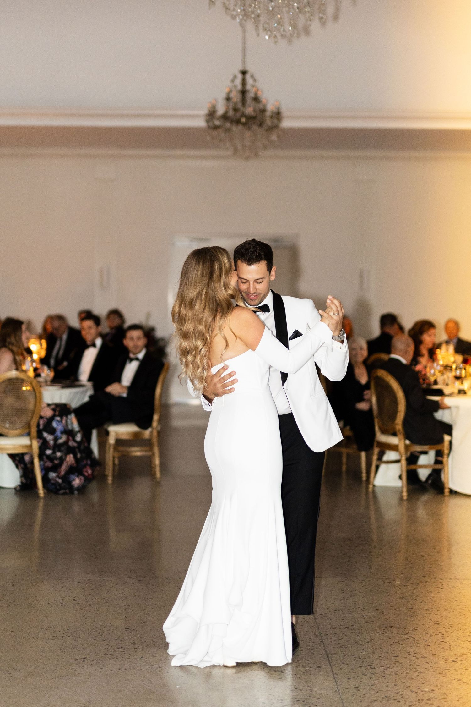 Couple dancing together in an elegant ballroom with chandeliers and wedding guests.