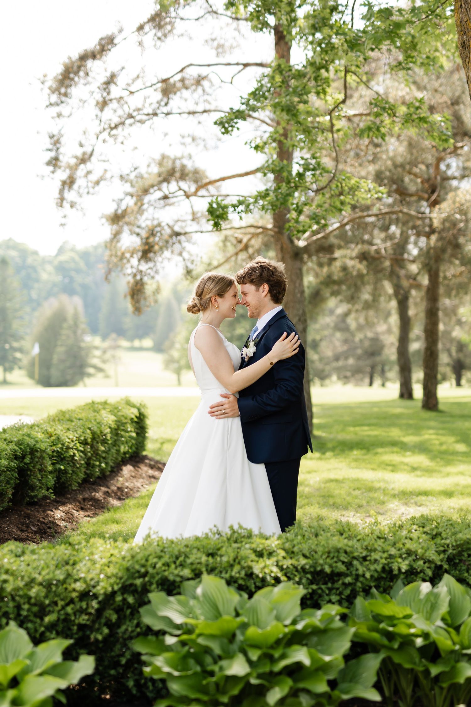 Bride and groom share intimate moment in garden setting with trees in background.
