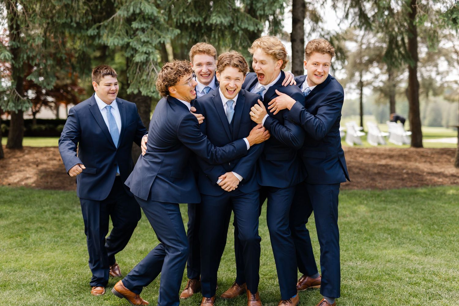 Groomsmen in navy suits pose playfully in a line on green grass.