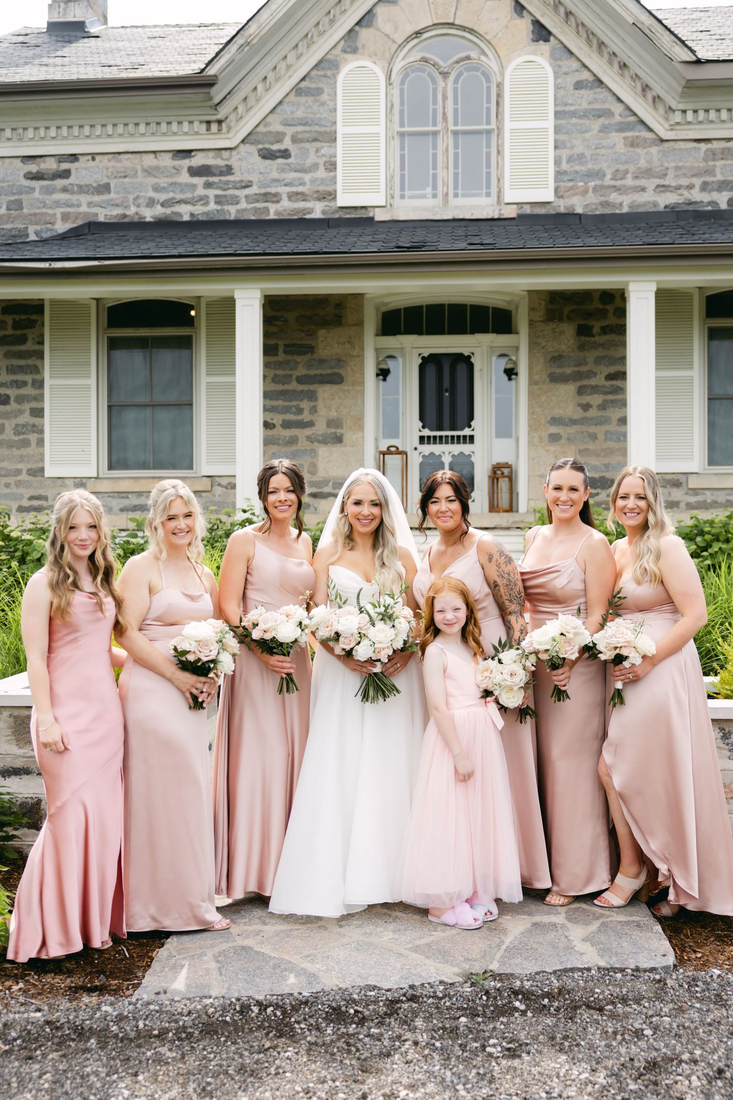 Bridal party wearing blush pink dresses pose in line against stone building facade.