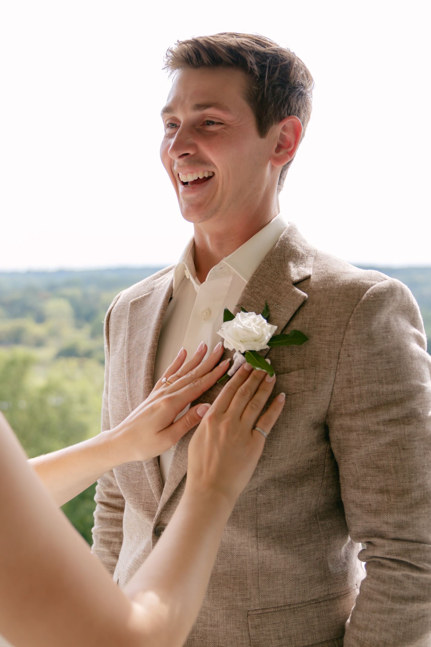 A boutonniere being pinned onto a tan suit jacket on a balcony with a scenic view.
