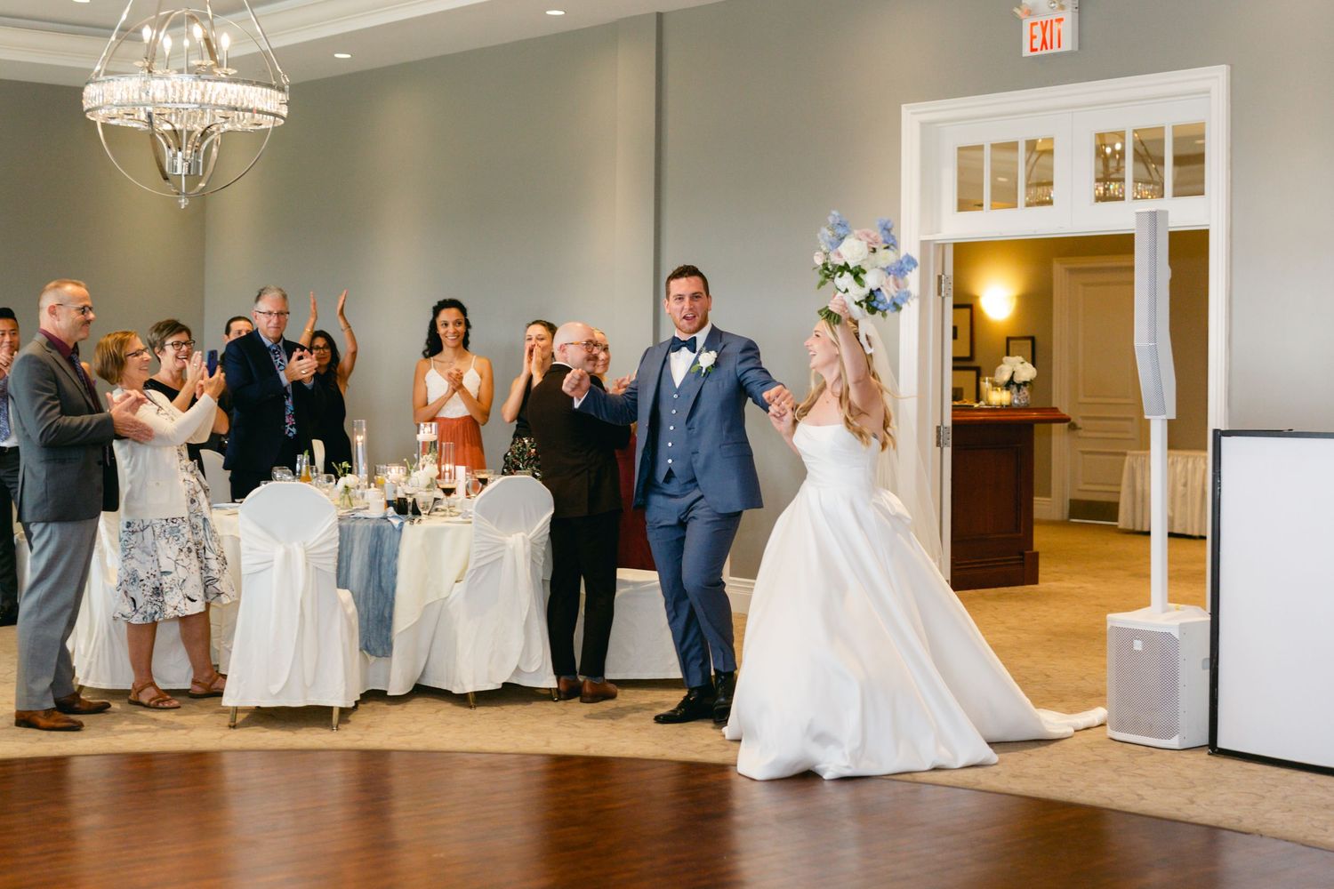 Wedding guests applaud during a celebration in an elegant ballroom with white chairs and a crystal chandelier.