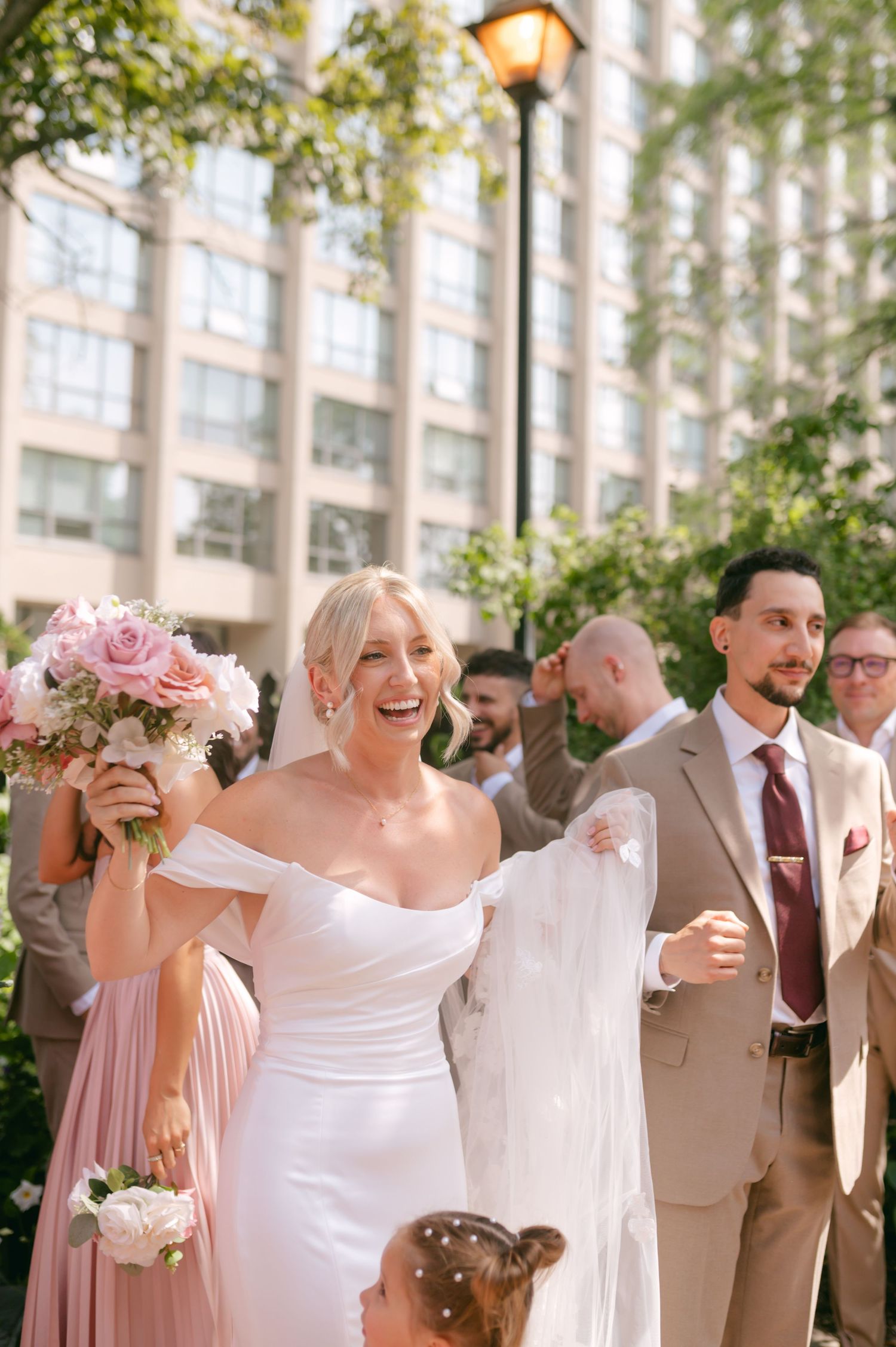 Wedding party celebrates outdoors with pink bouquets and tan suits near a vintage lamppost.