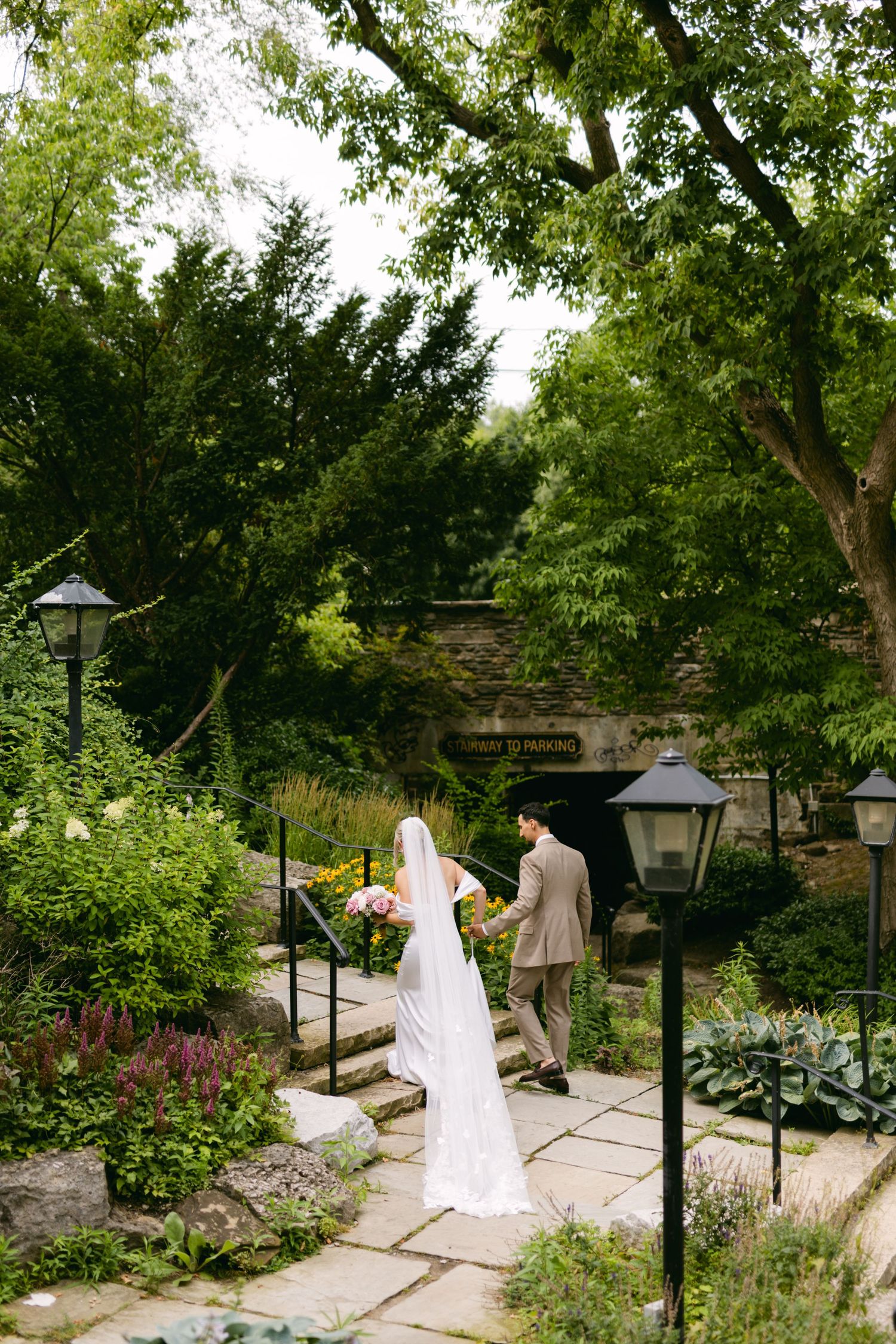 Wedding couple walks through a garden path lined with lamp posts and blooming flowers.