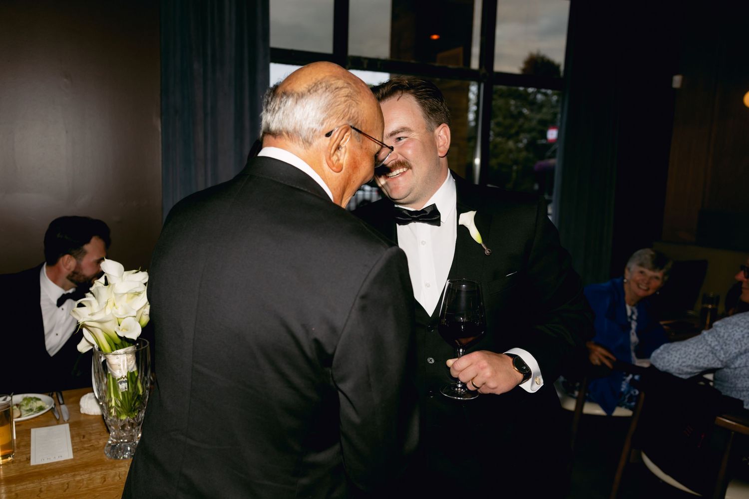Two men in tuxedos share a conversation at a wedding reception while holding drinks.