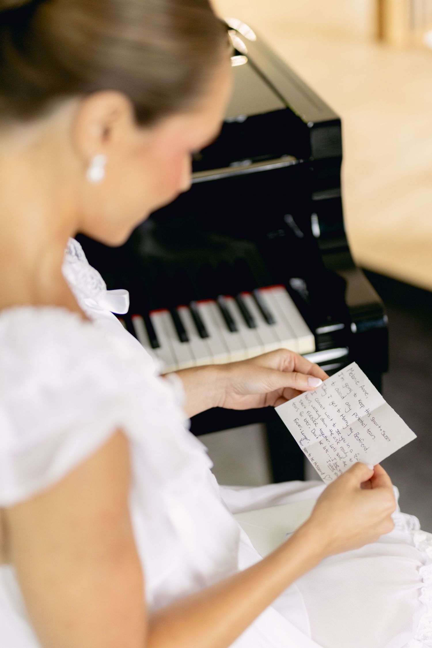 Bride in white dress reading handwritten letter while seated at black grand piano.