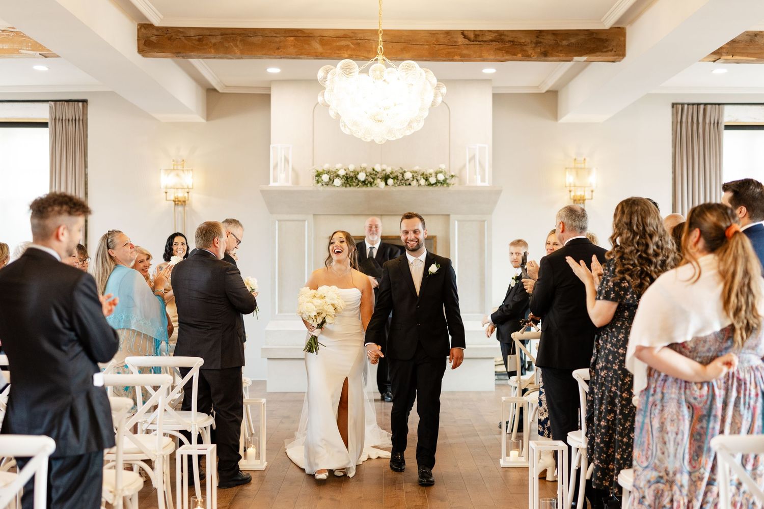 Bride and groom walk back down aisle after ceremony as guests applaud in elegant white venue.