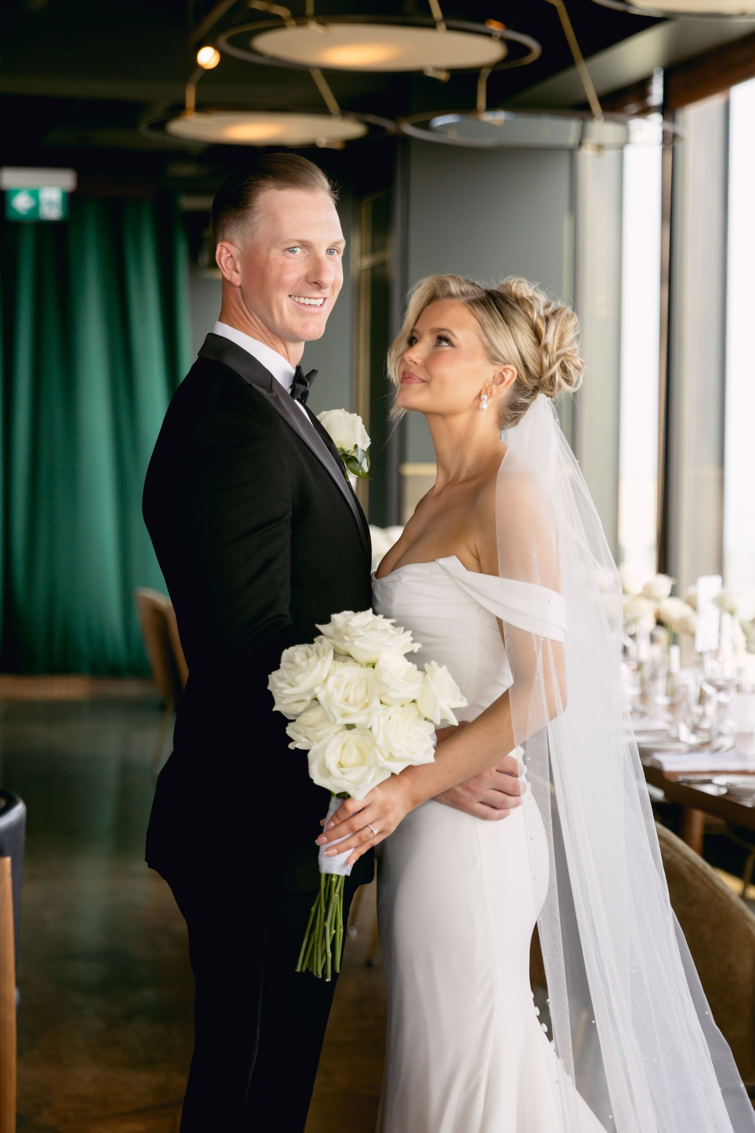 Smiling groom in black tuxedo holds bride in white off-shoulder gown with white rose bouquet indoors.