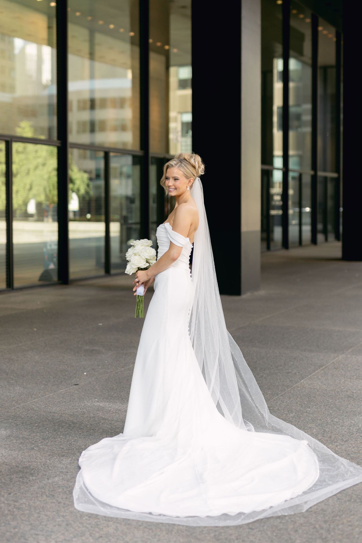 Bride in fitted white off-shoulder gown with long veil holds white bouquet outside glass building.