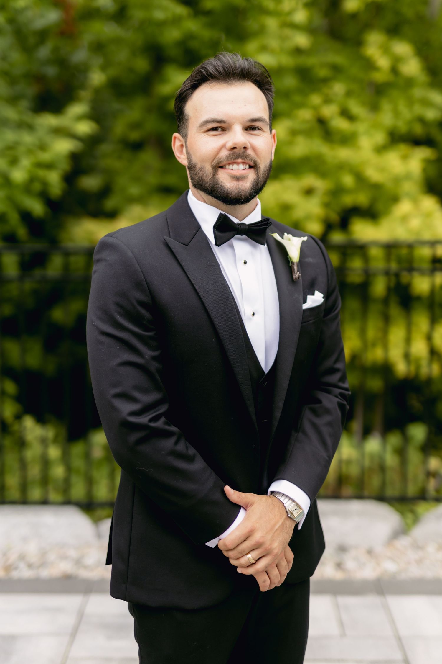 Handsome groom in black tuxedo with bow tie and white boutonniere posing outdoors.