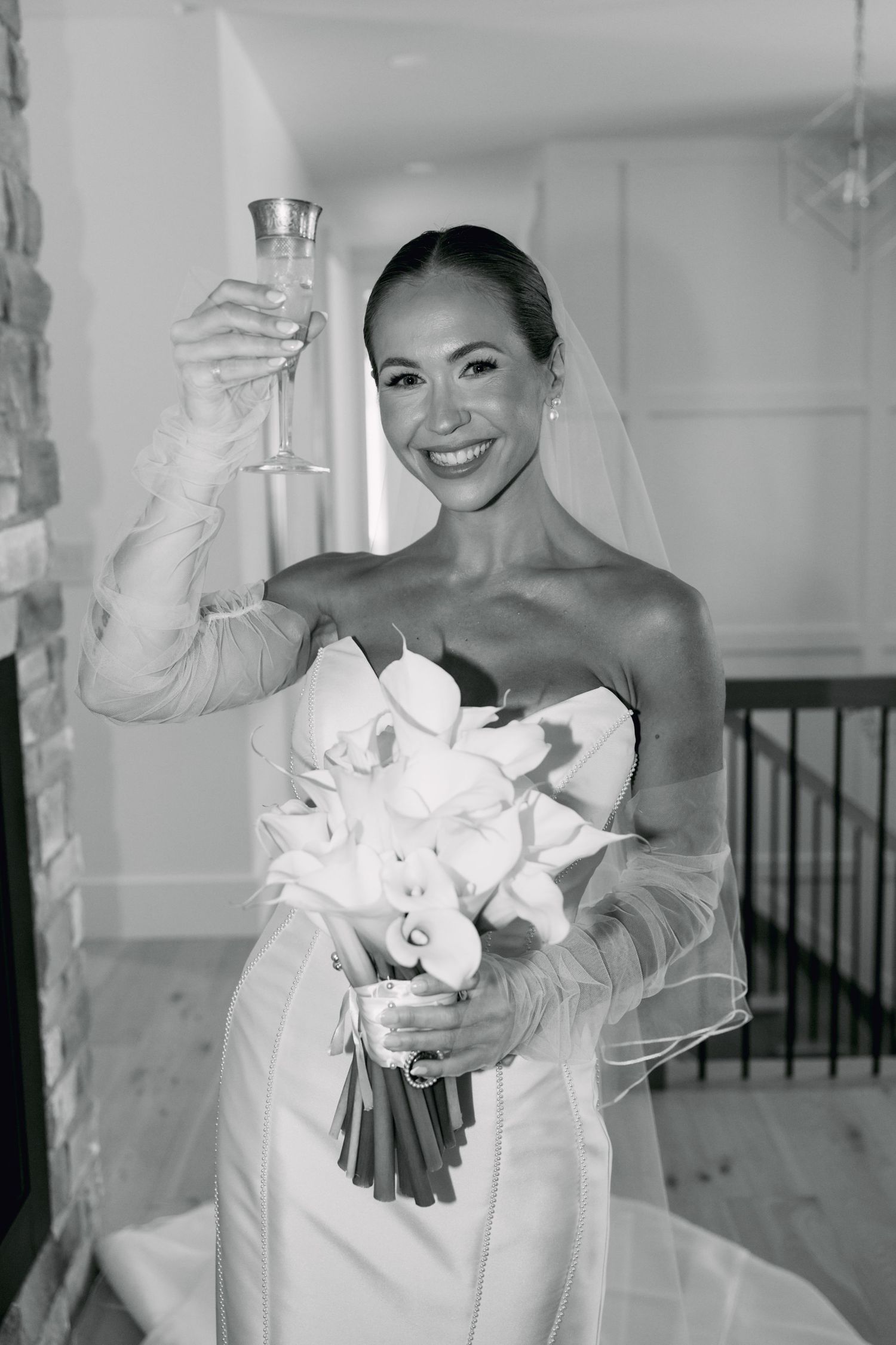 Smiling bride in strapless gown raises champagne glass while holding white calla lily bouquet in black and white photo.