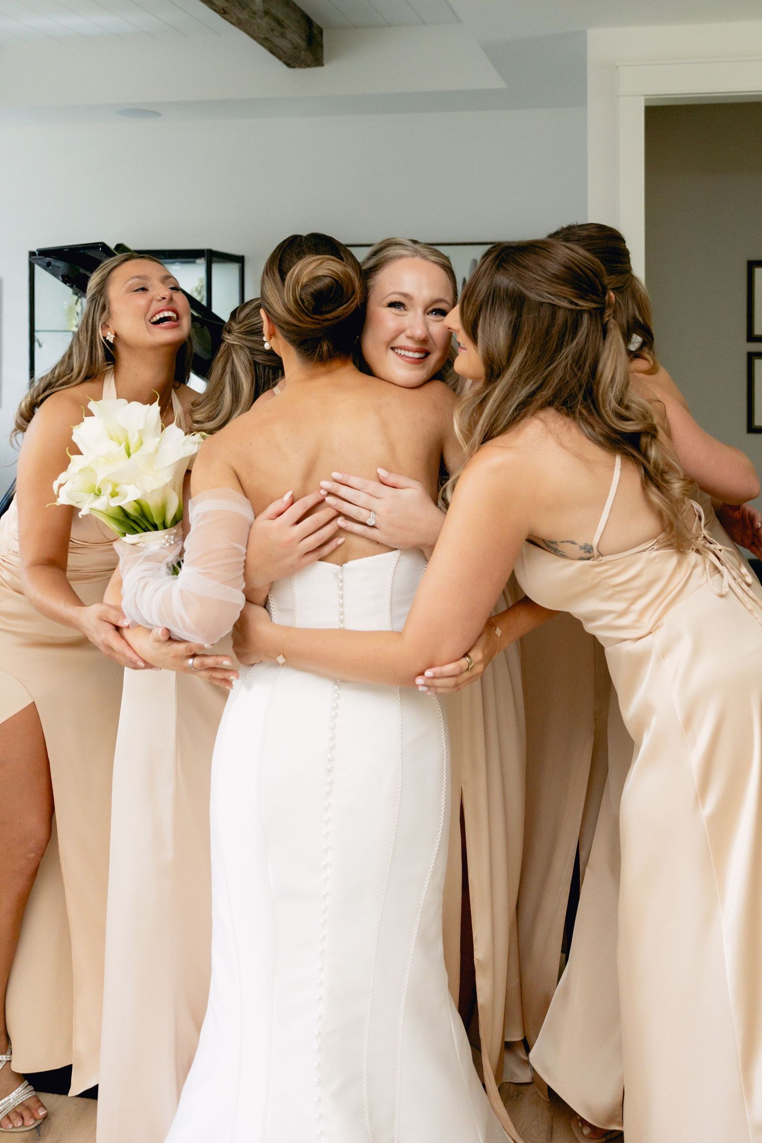 Bride in white gown embraced by smiling bridesmaids in champagne dresses holding white calla lily bouquet.