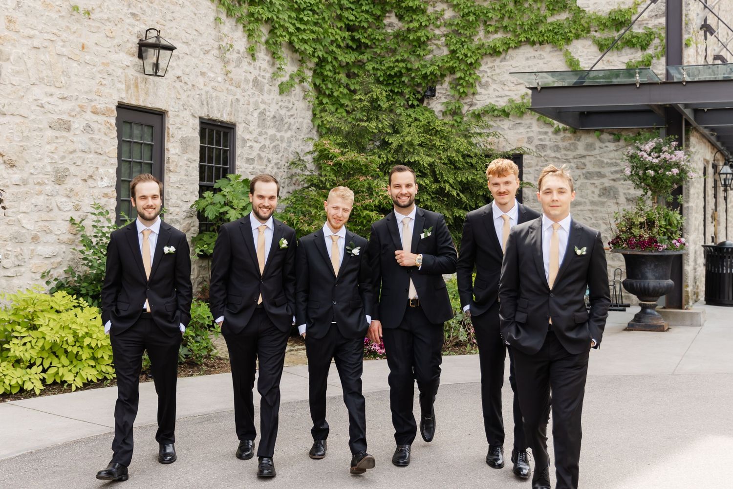 Six groomsmen in black suits with white ties pose outdoors near a stone building covered in ivy.