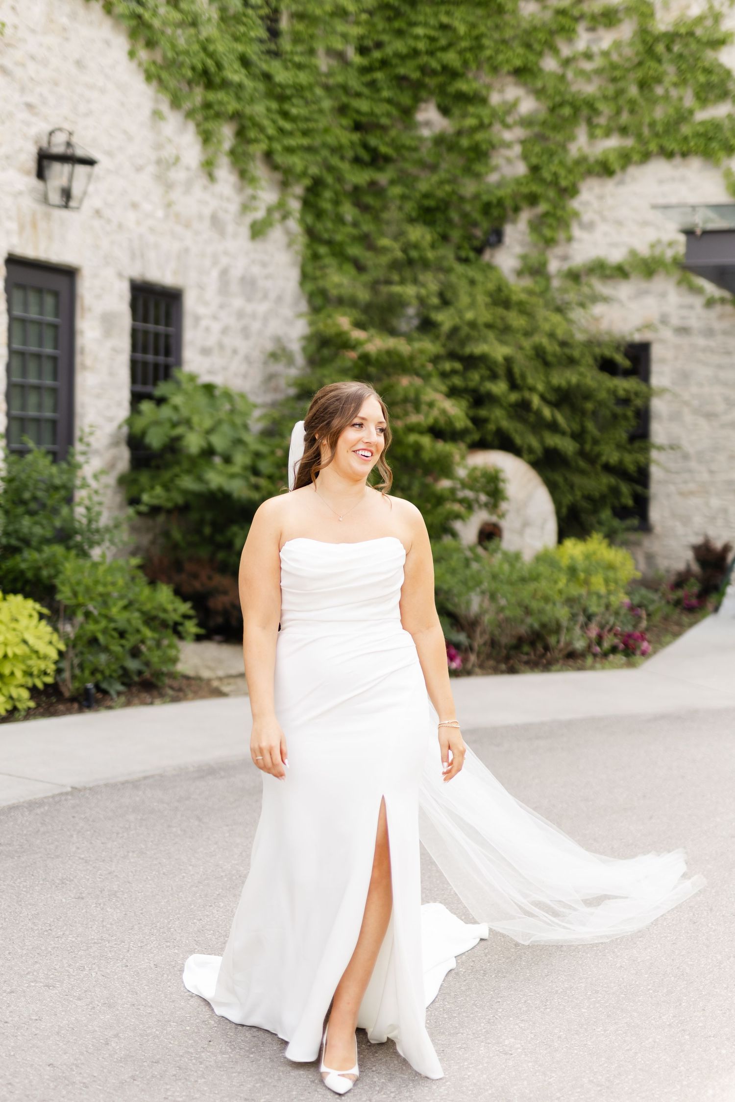 Radiant bride in a strapless white fitted wedding dress with a slit twirls outdoors at a stone venue.