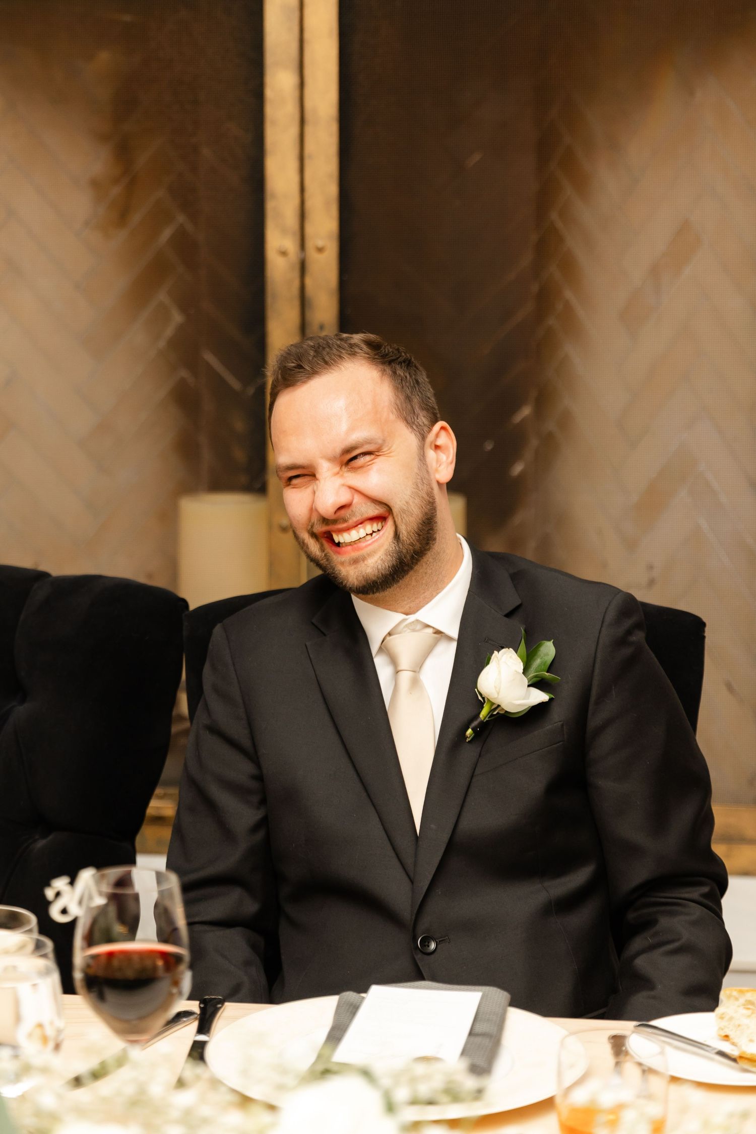 Smiling groom in a black suit with white boutonniere laughs joyfully at the wedding reception dinner table.