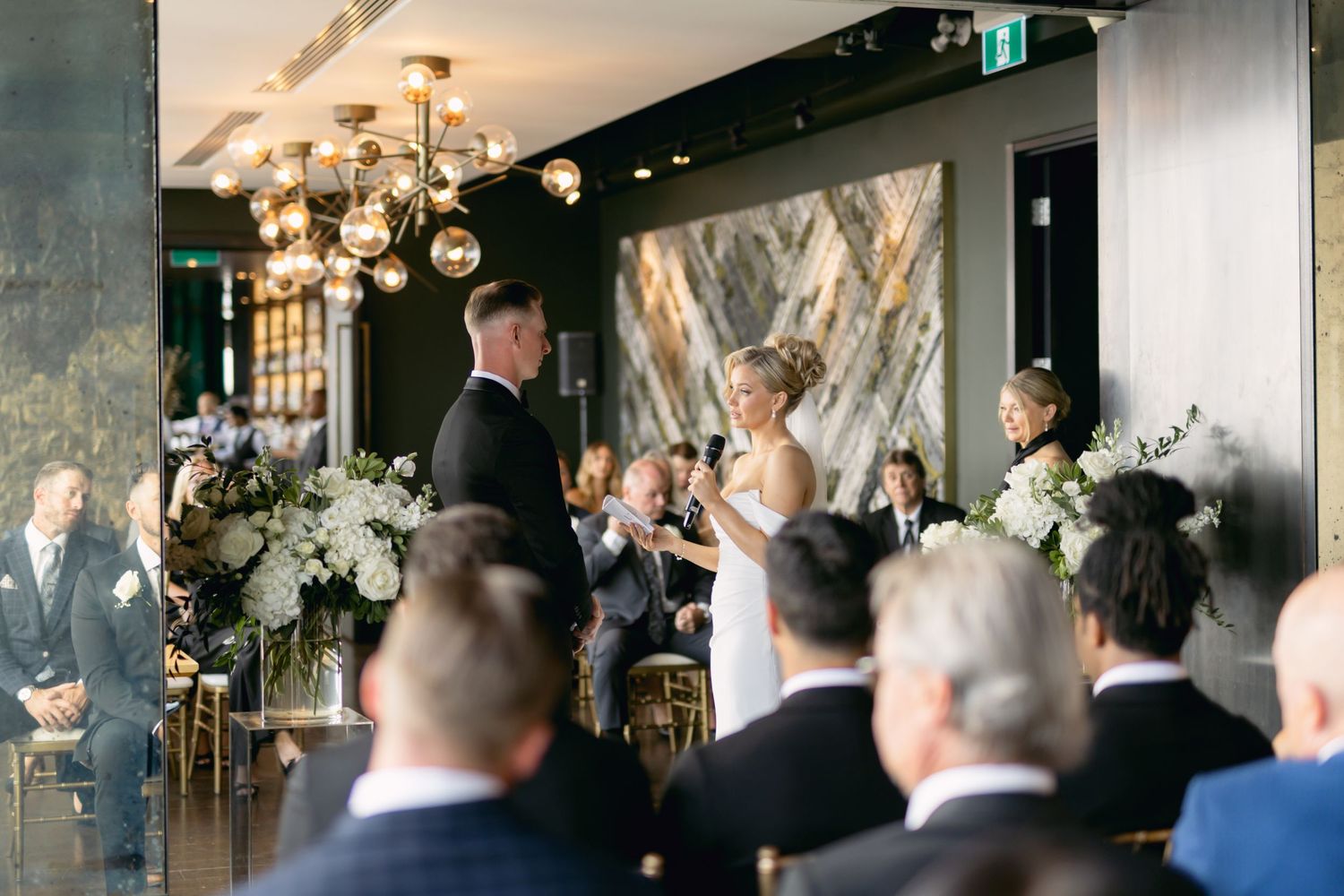 Elegant wedding ceremony in a modern venue with floral arrangements, guests seated, and couple toasting under golden chandelier.