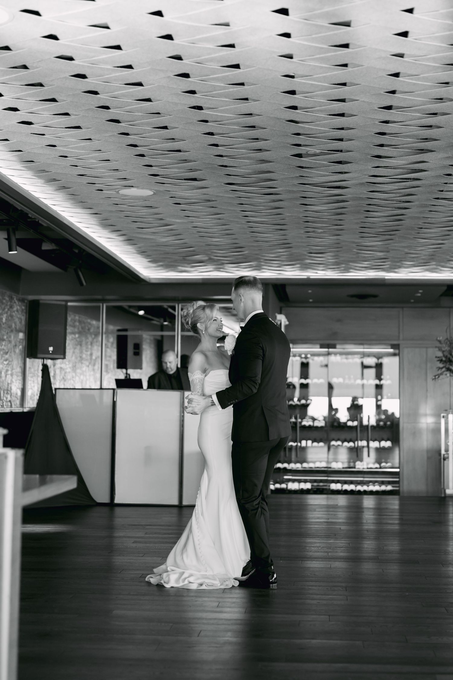 Black and white photo of bride and groom sharing first dance in elegant reception venue with patterned ceiling.