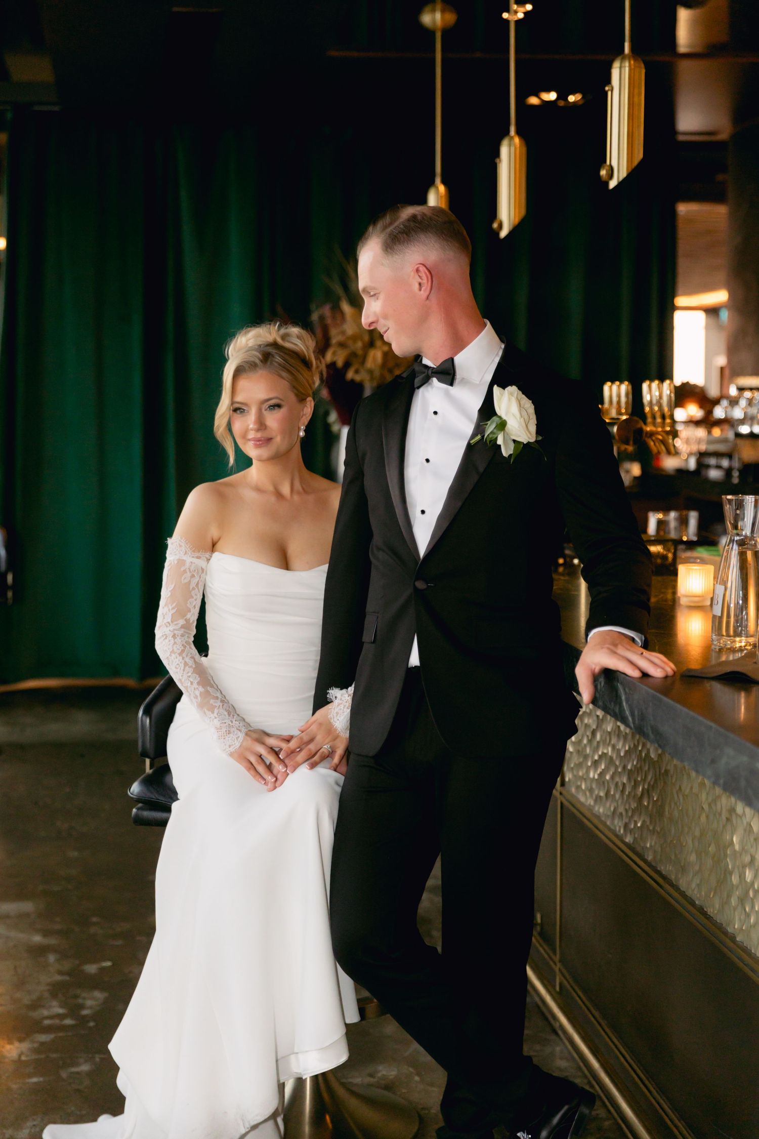 Bride in off-shoulder lace gown and groom in black tuxedo pose together at a moody green bar.