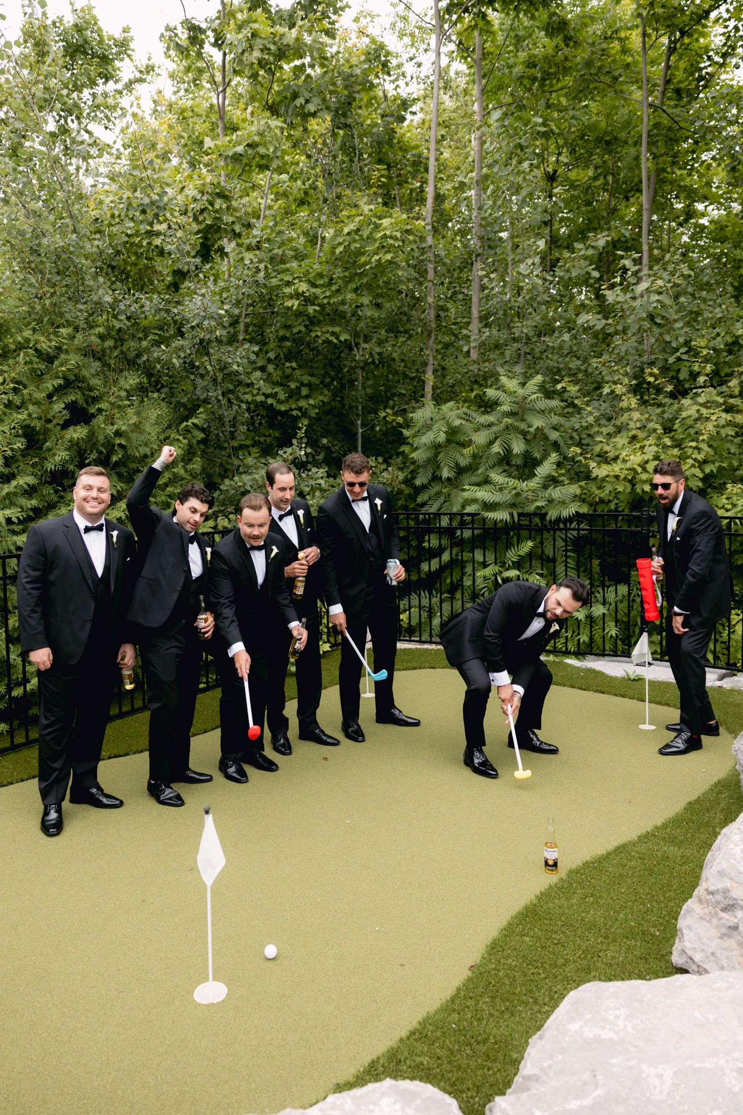 Groomsmen in black tuxedos celebrate on a putting green, cheering as one golfs before the wedding.