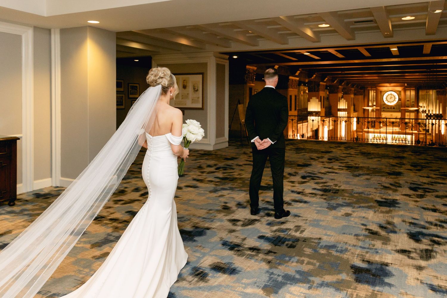 Bride in elegant backless gown with long veil approaches groom in black suit for first look in hotel ballroom.