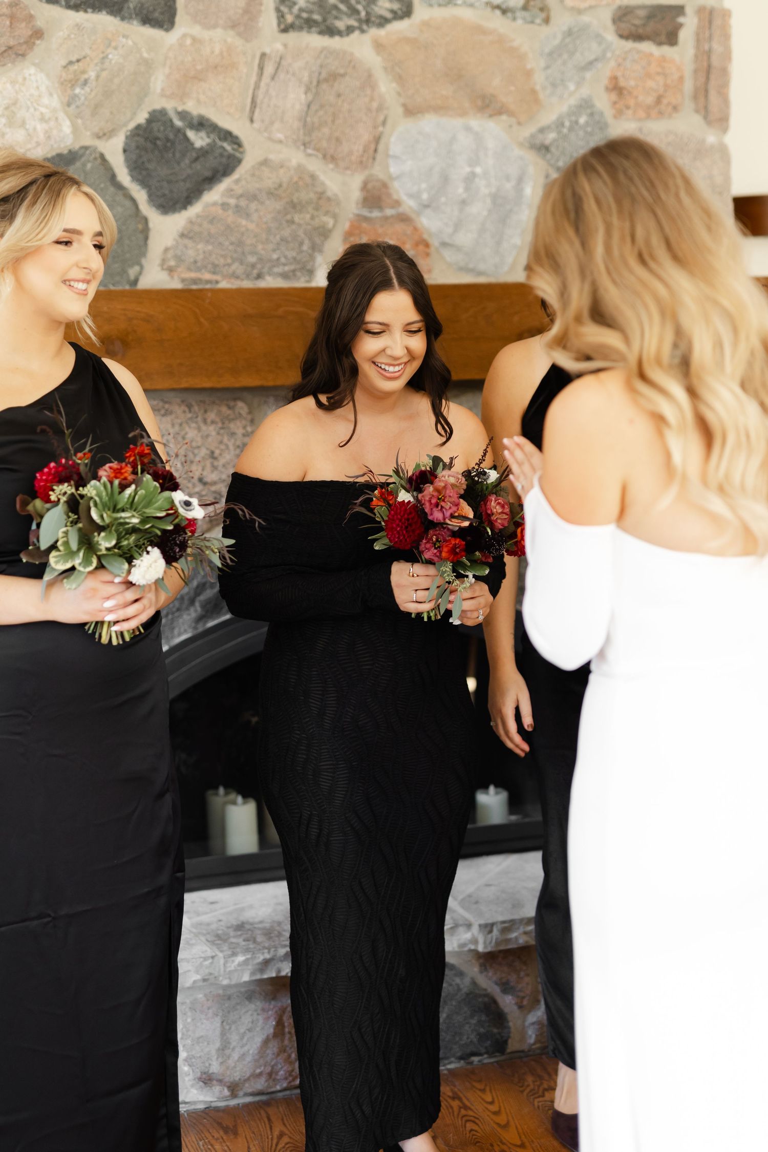 Bridesmaids in black dresses gather around bride in white gown against stone wall.