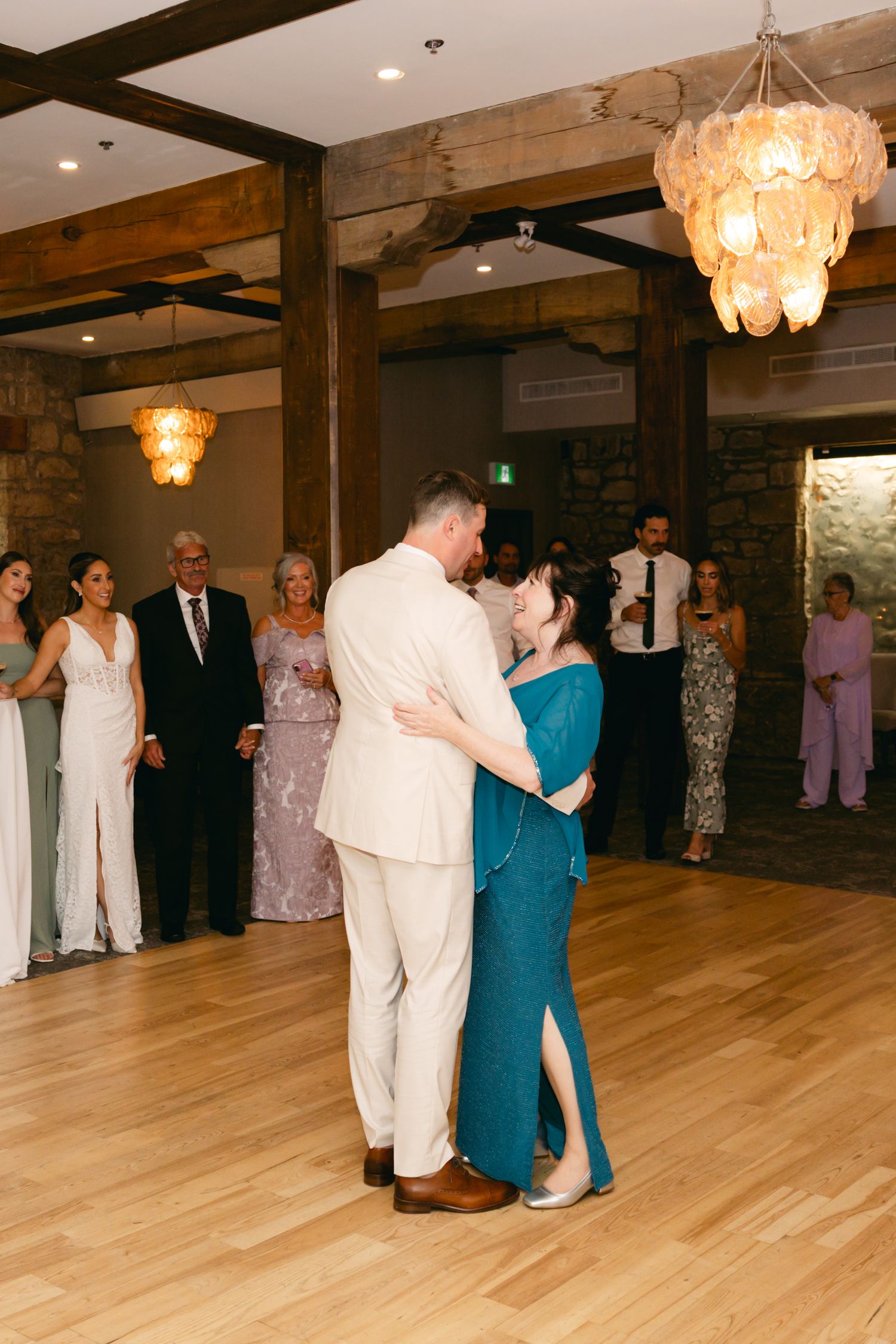 Wedding guests dancing in formal attire on a wooden dance floor under crystal chandeliers.