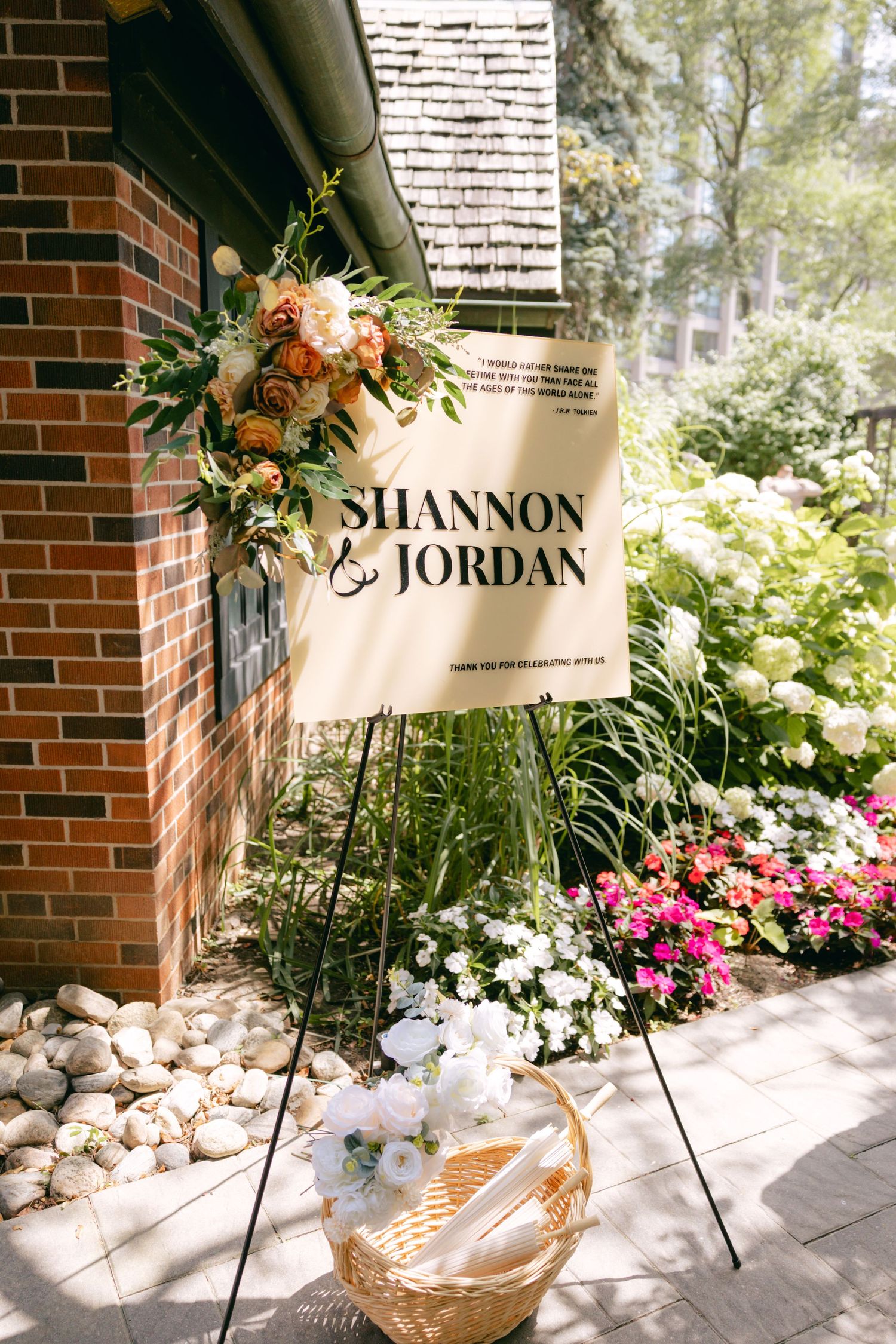 A wedding welcome sign decorated with peach and white flowers against a brick wall.