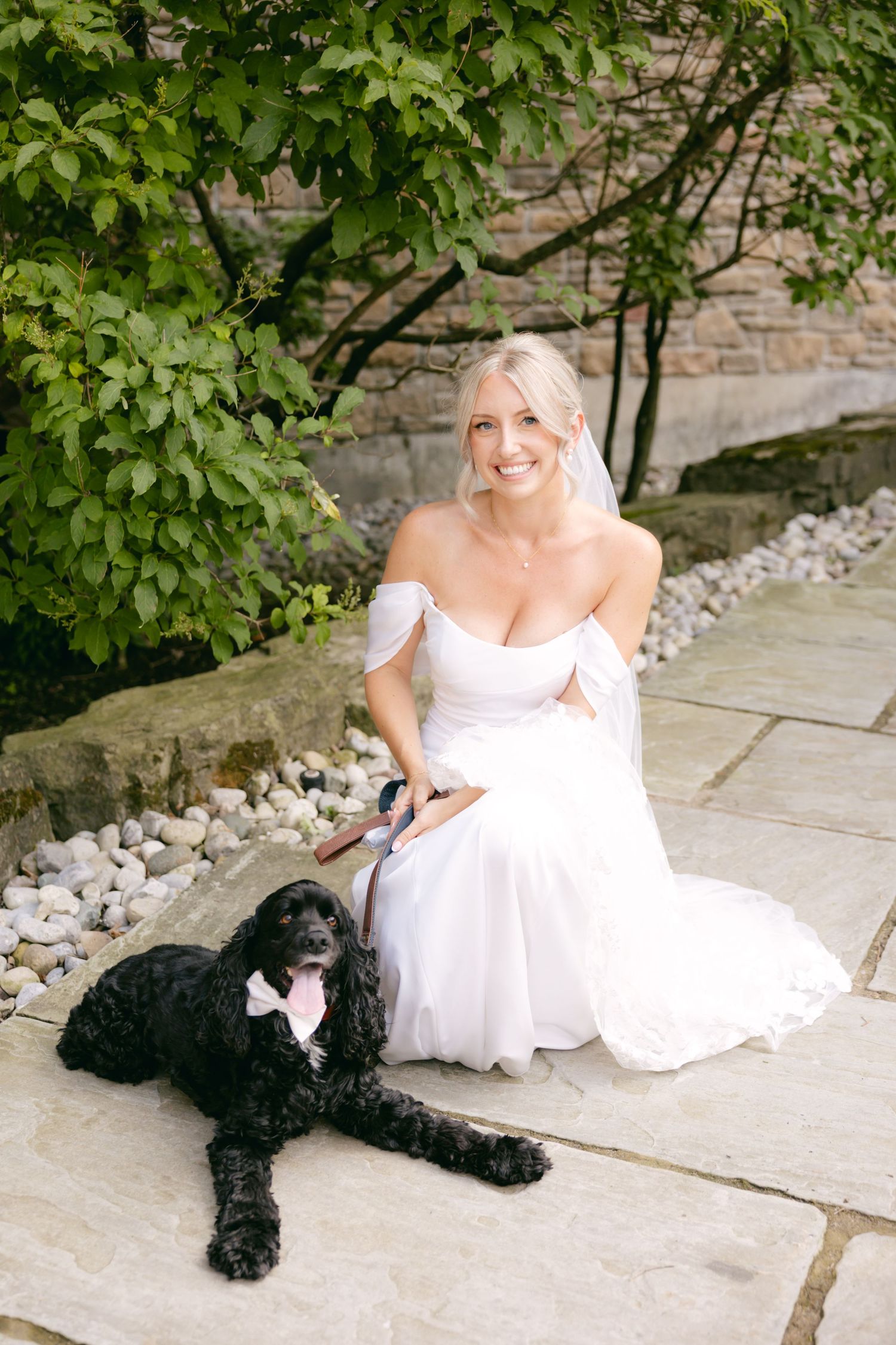 A bride in white dress sits with her black dog on stone steps surrounded by greenery.