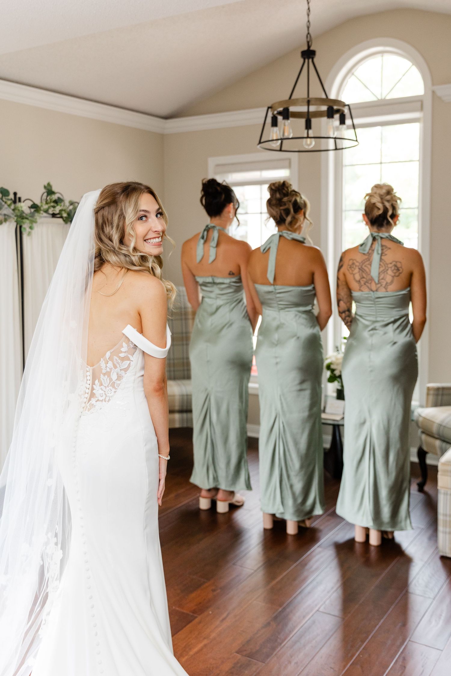 Bridesmaids in matching sage green dresses pose with the bride in her lace wedding gown.